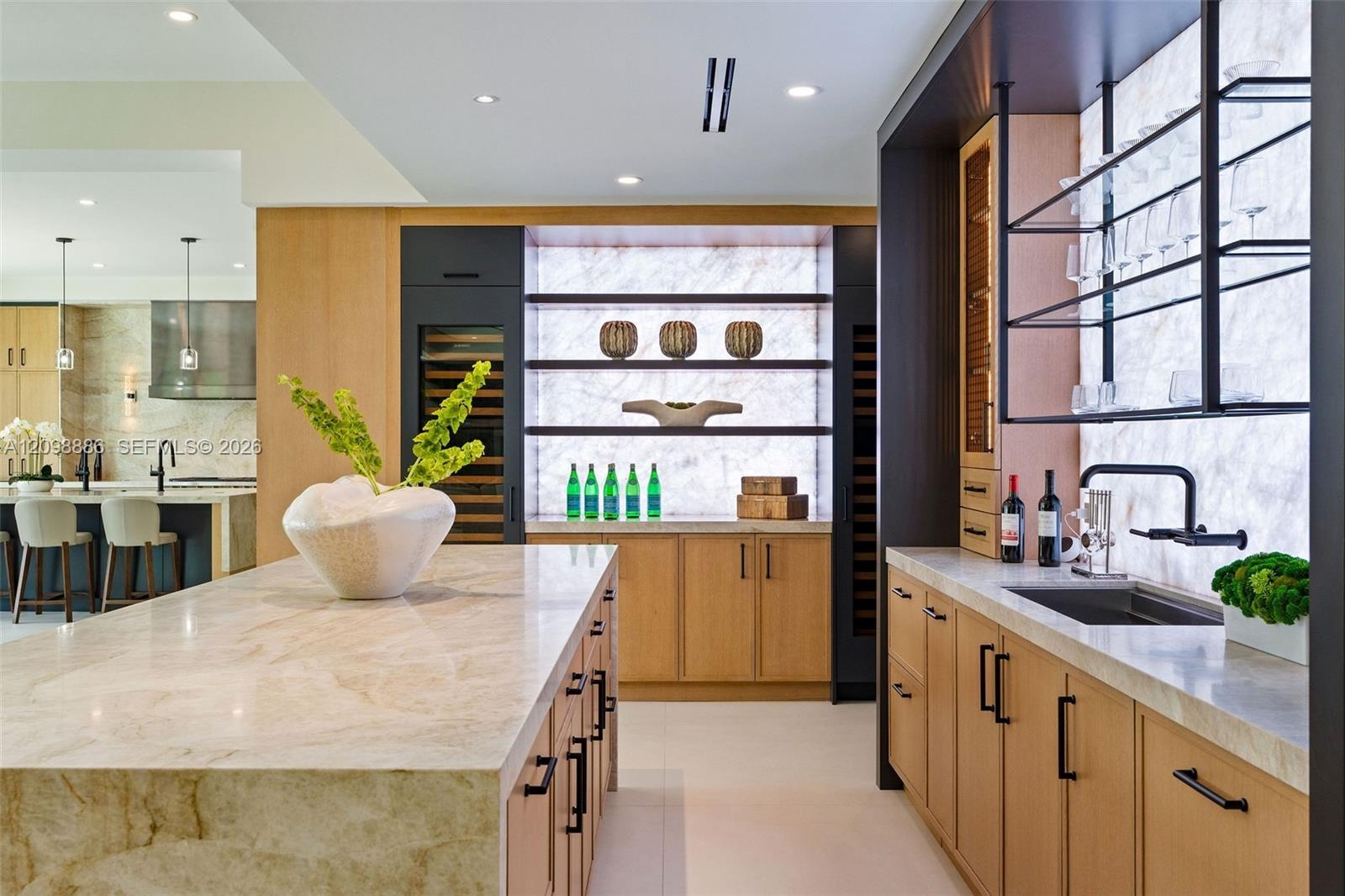 Custom kitchen featuring a large stone-surfaced island, light wood cabinetry, integrated wine refrigeration, an illuminated stone backsplash, and matt