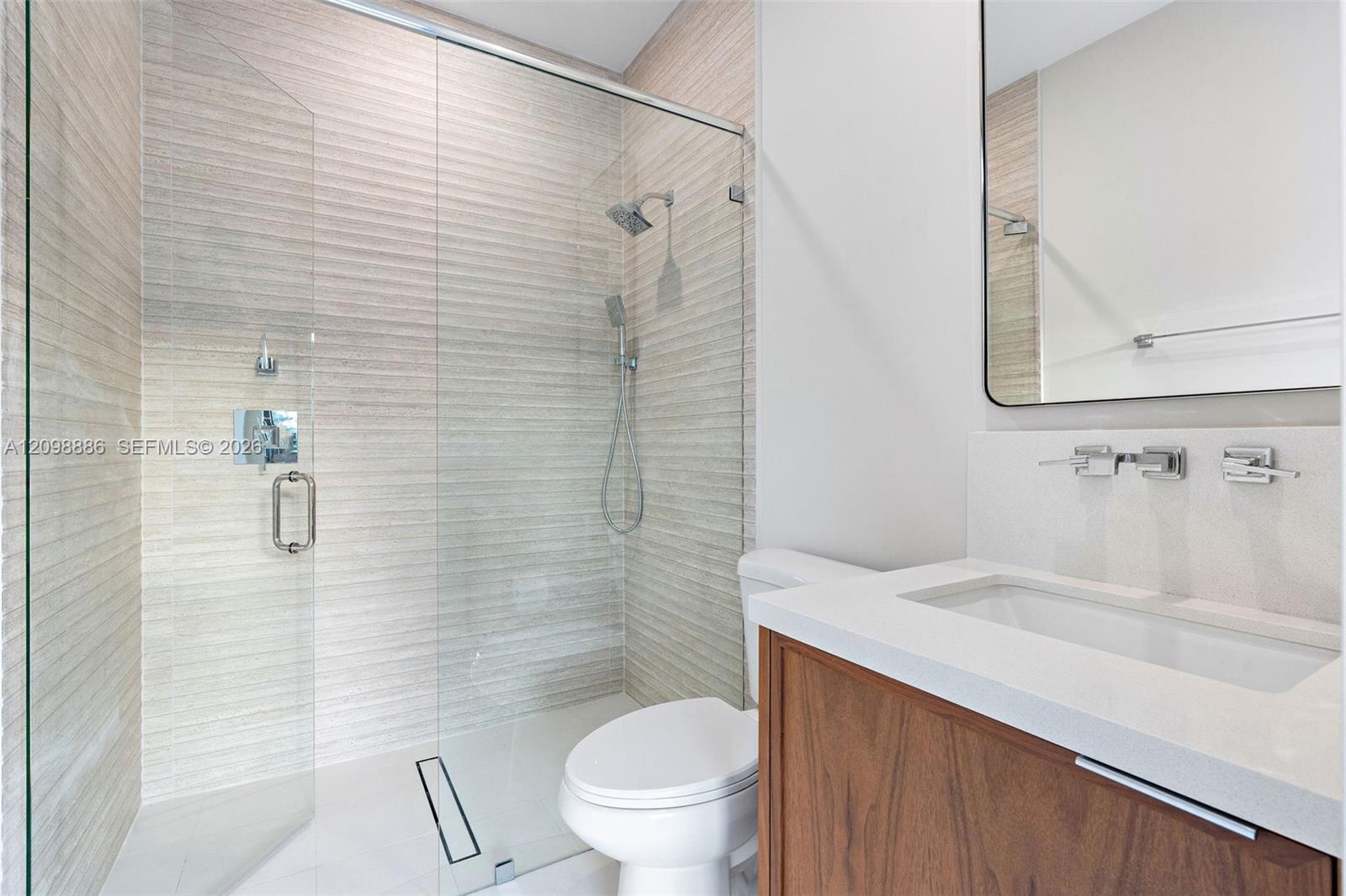 Walk-in shower featuring textured wall tile, glass enclosure, and rainfall showerhead, paired with a wood-finish vanity, solid-surface countertop, and