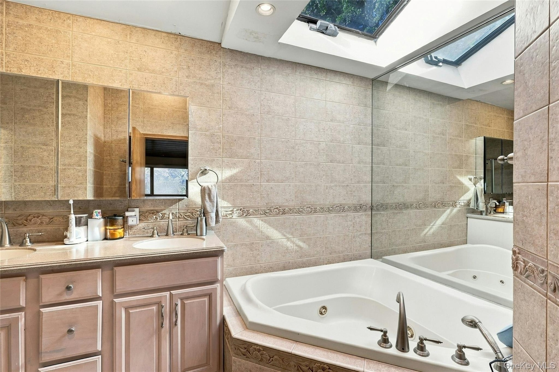 Custom wooden vanity with dual recessed-mirror cabinets, corner jetted tub, wall-to-wall tiling with decorative trim, and an expansive overhead skylig