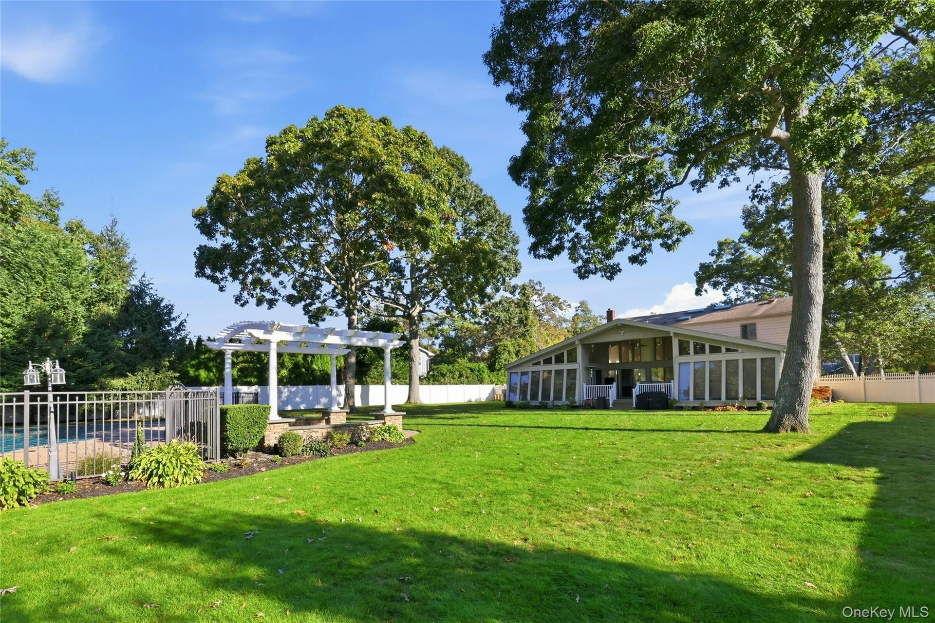 Expansive backyard featuring a sprawling lawn, mature trees, an inground pool with metal perimeter fencing, and a white architectural pergola
