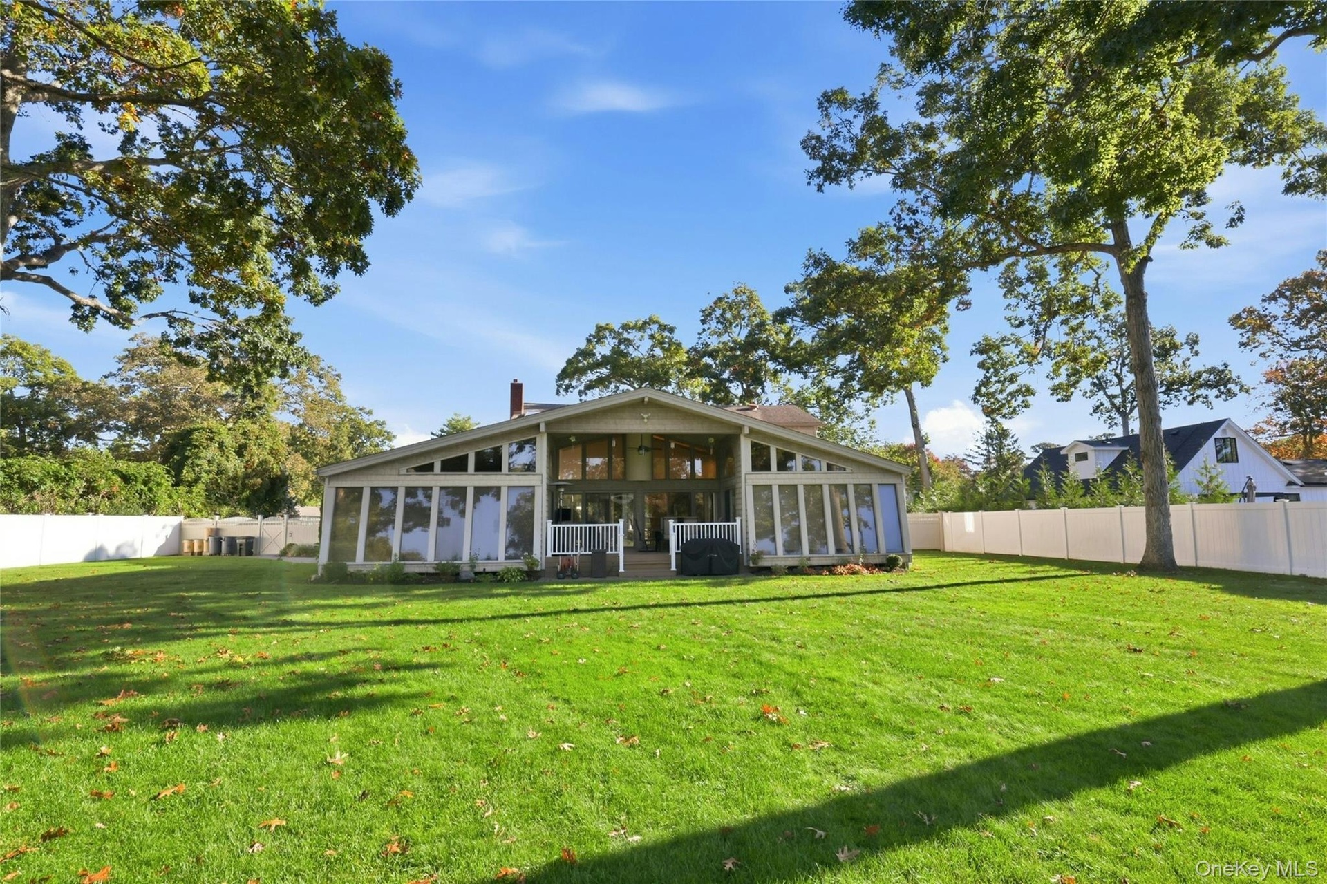 Expansive rear yard featuring a manicured lawn, perimeter white privacy fencing, mature trees, and a multi-windowed architectural exterior with a rais