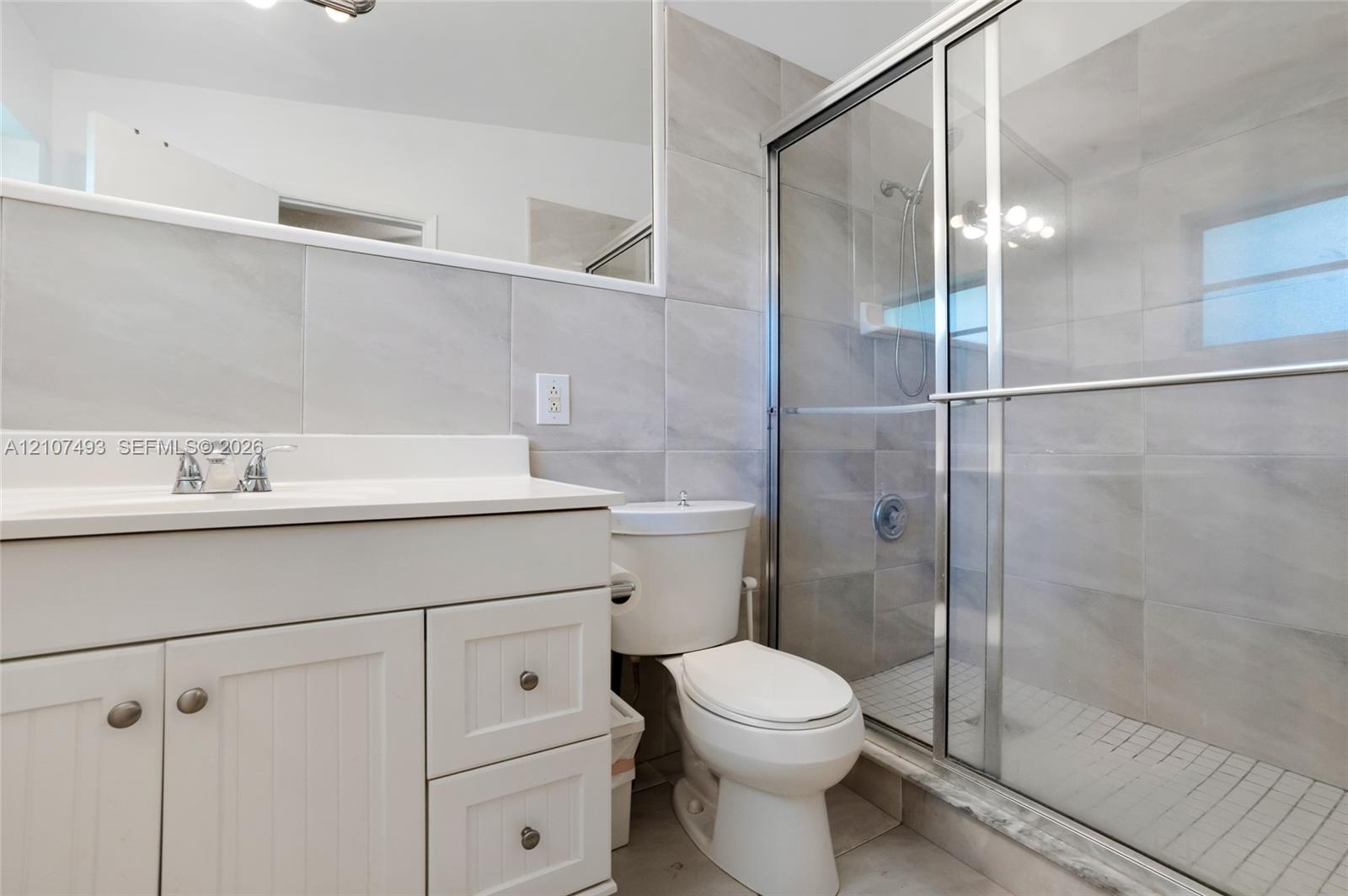 Bathroom featuring a white vanity cabinet, large wall-mounted mirror, walk-in shower with sliding glass doors, and light-toned tile wall finishes
