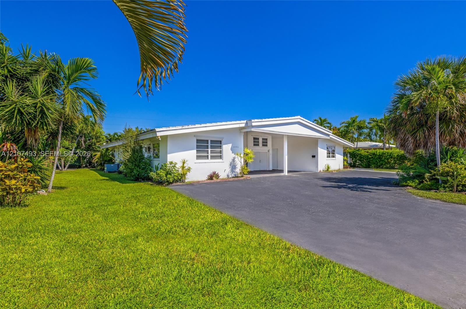 Single-story residence featuring white exterior cladding, a covered carport, and asphalt driveway, surrounded by mature palm trees and landscaped grou