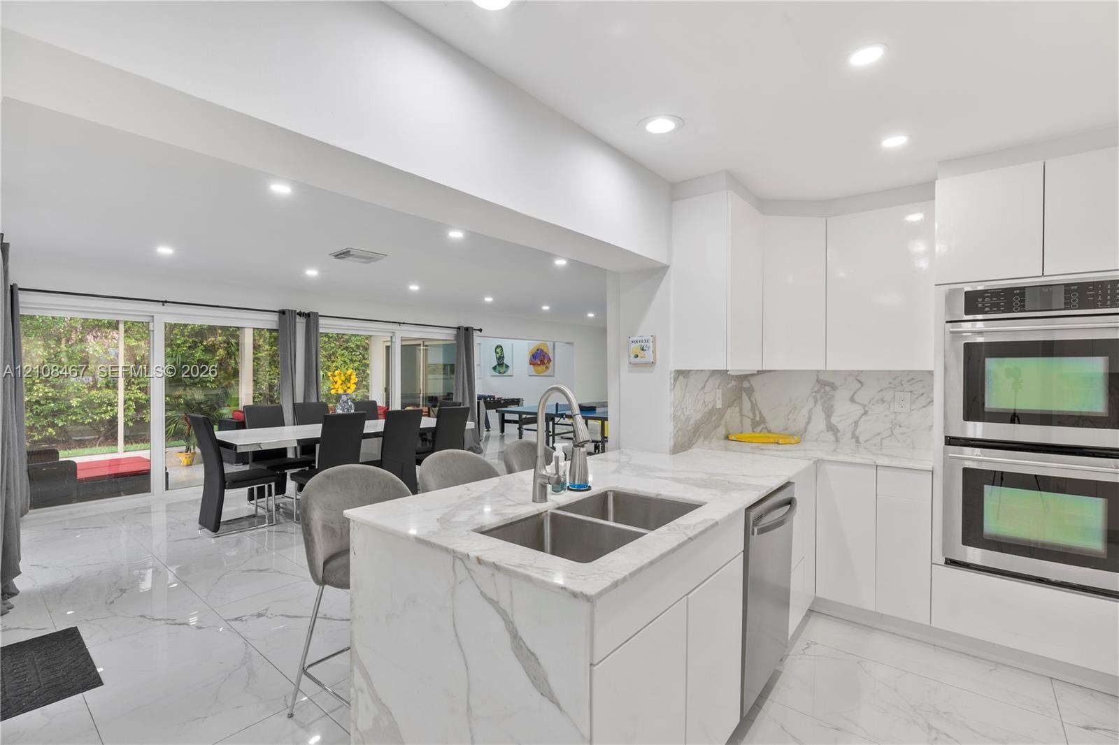 Kitchen featuring white flat-panel cabinetry, stone-patterned countertops, a central island with a double-basin stainless steel sink, built-in double 