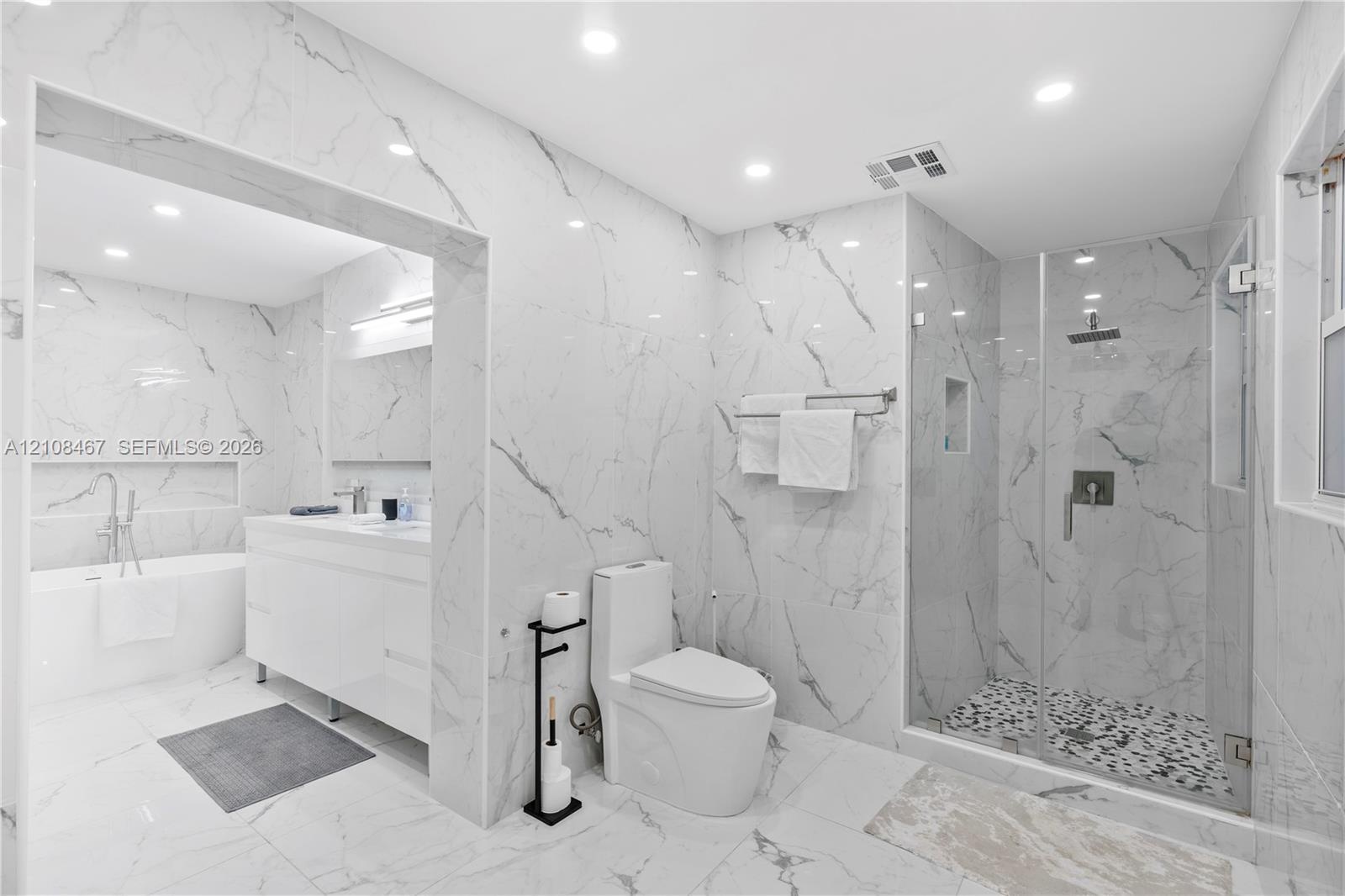 Full-height wall-to-wall stone-finish tile, glass-enclosed shower with pebble-finish flooring, built-in recessed wall niche, freestanding soaking tub,