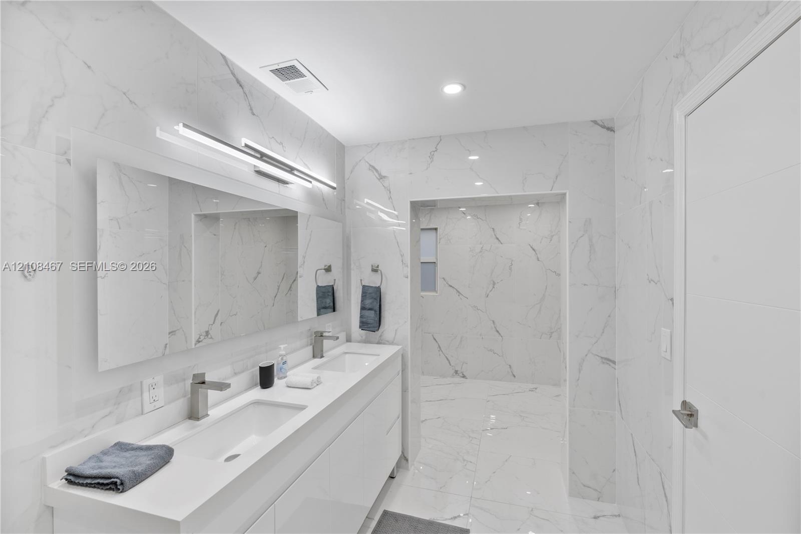 Dual-sink vanity with white countertop, full-width mirror, integrated wall-mounted lighting, floor-to-ceiling stone-look wall tiling, and walk-in show