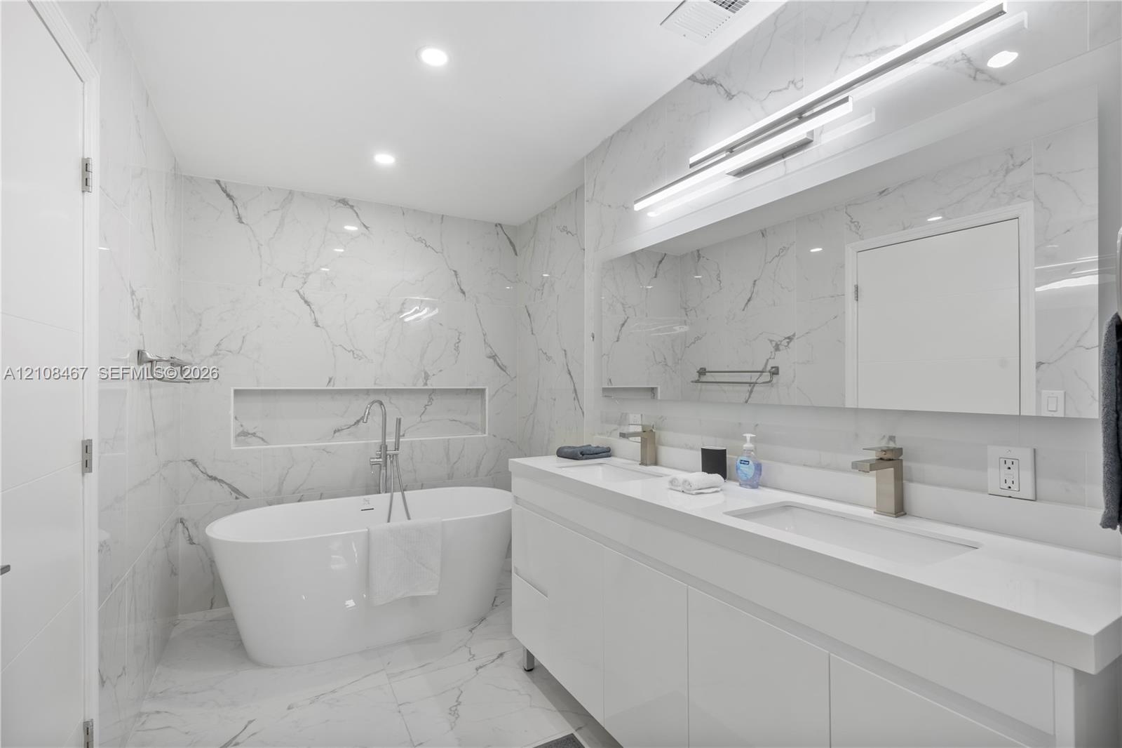 Modern bathroom featuring floor-to-ceiling stone-look tiling, a freestanding soaking tub with a wall-recessed niche, a dual-sink floating vanity, inte