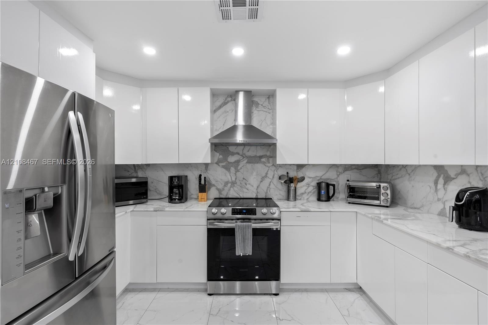Modern kitchen featuring high-gloss white cabinetry, stone-surface countertops, full-height stone backsplash, stainless steel appliances, and large-fo