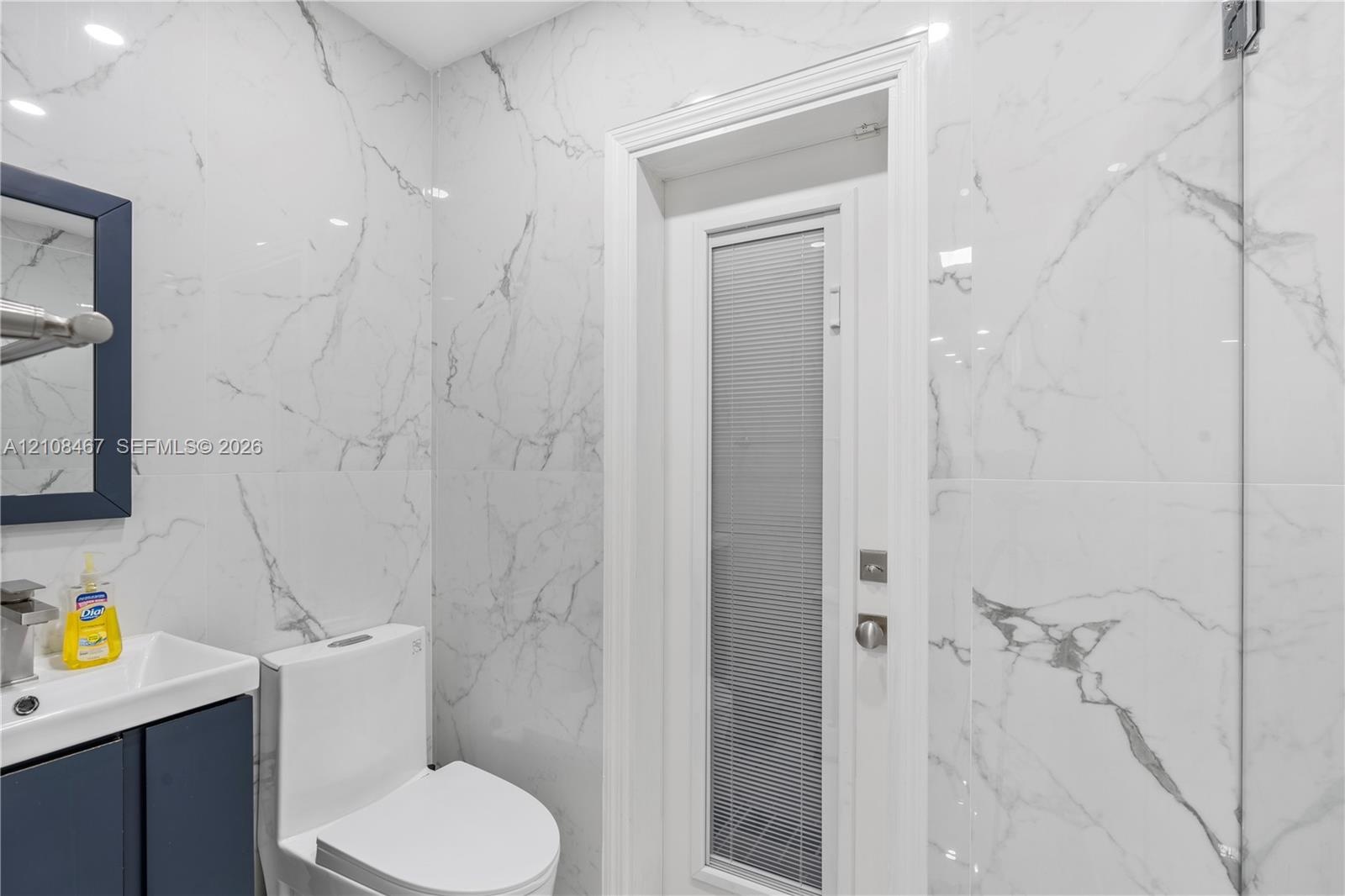 Full-height wall tiling with a veined pattern, vanity featuring a rectangular integrated sink, solid-color cabinetry, and a door with an integrated pr