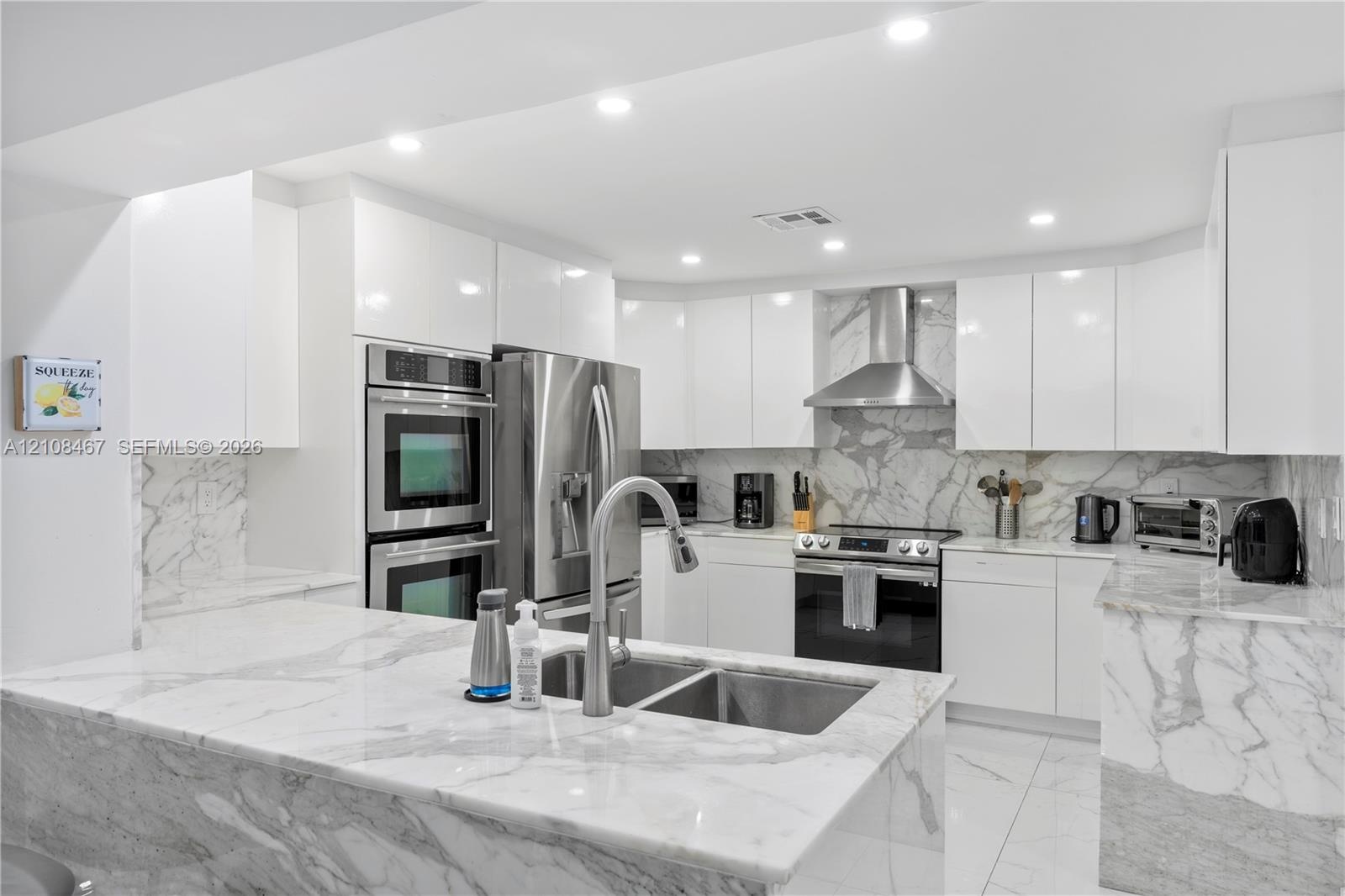 Modern kitchen featuring high-gloss white cabinetry, stone countertops and backsplash, stainless steel built-in double ovens, stainless steel range ho