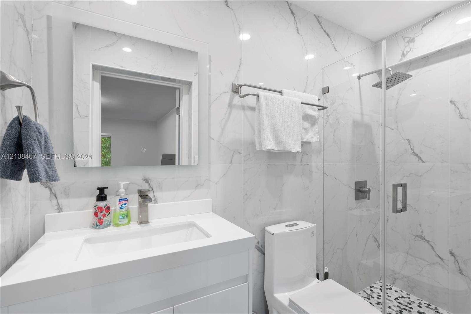 Bathroom featuring large-format wall tiling with veining detail, glass-enclosed shower with a rainfall showerhead, vanity with integrated basin, and m