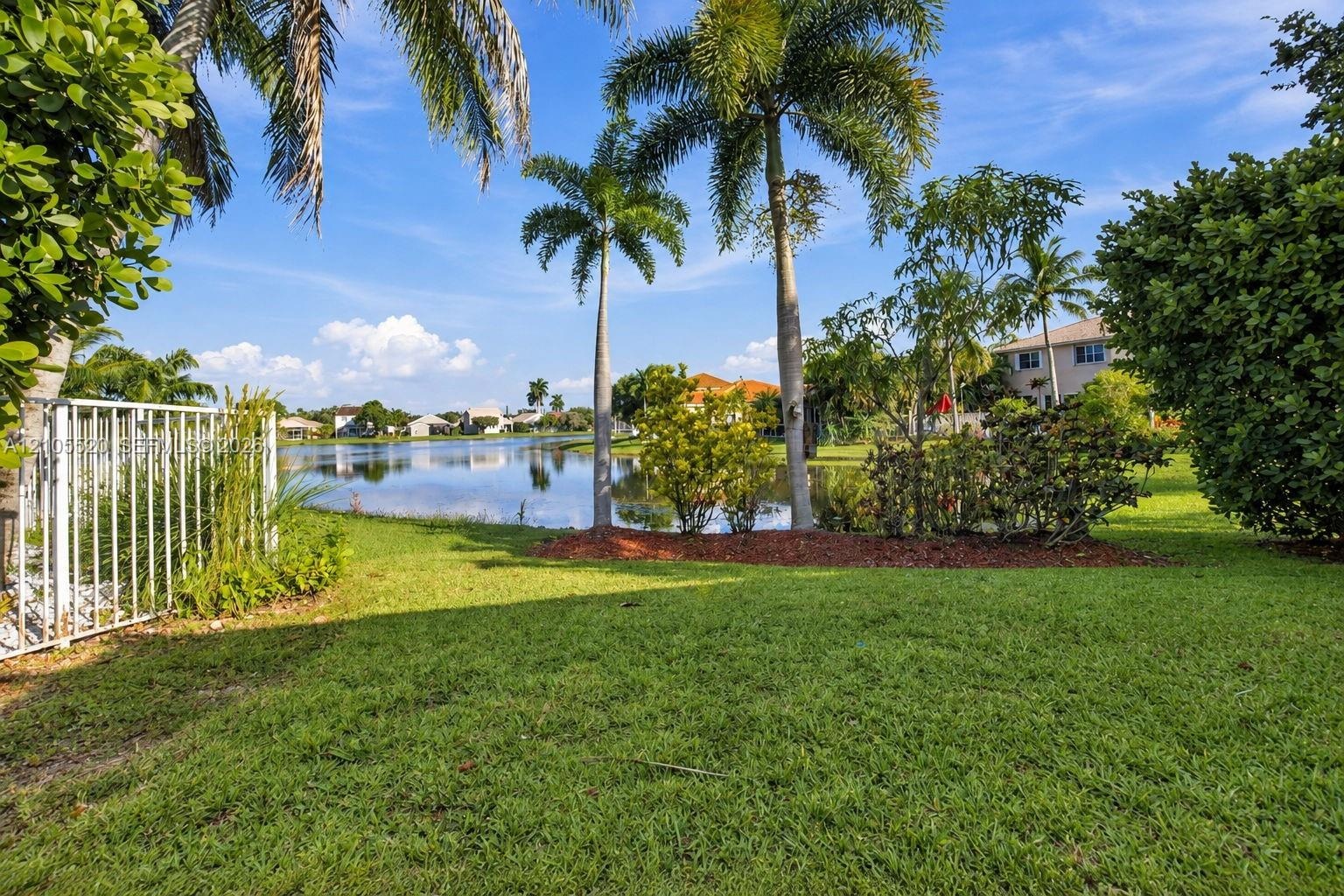 Waterfront backyard featuring a manicured lawn, established palm trees, decorative mulch beds, and white metal perimeter fencing