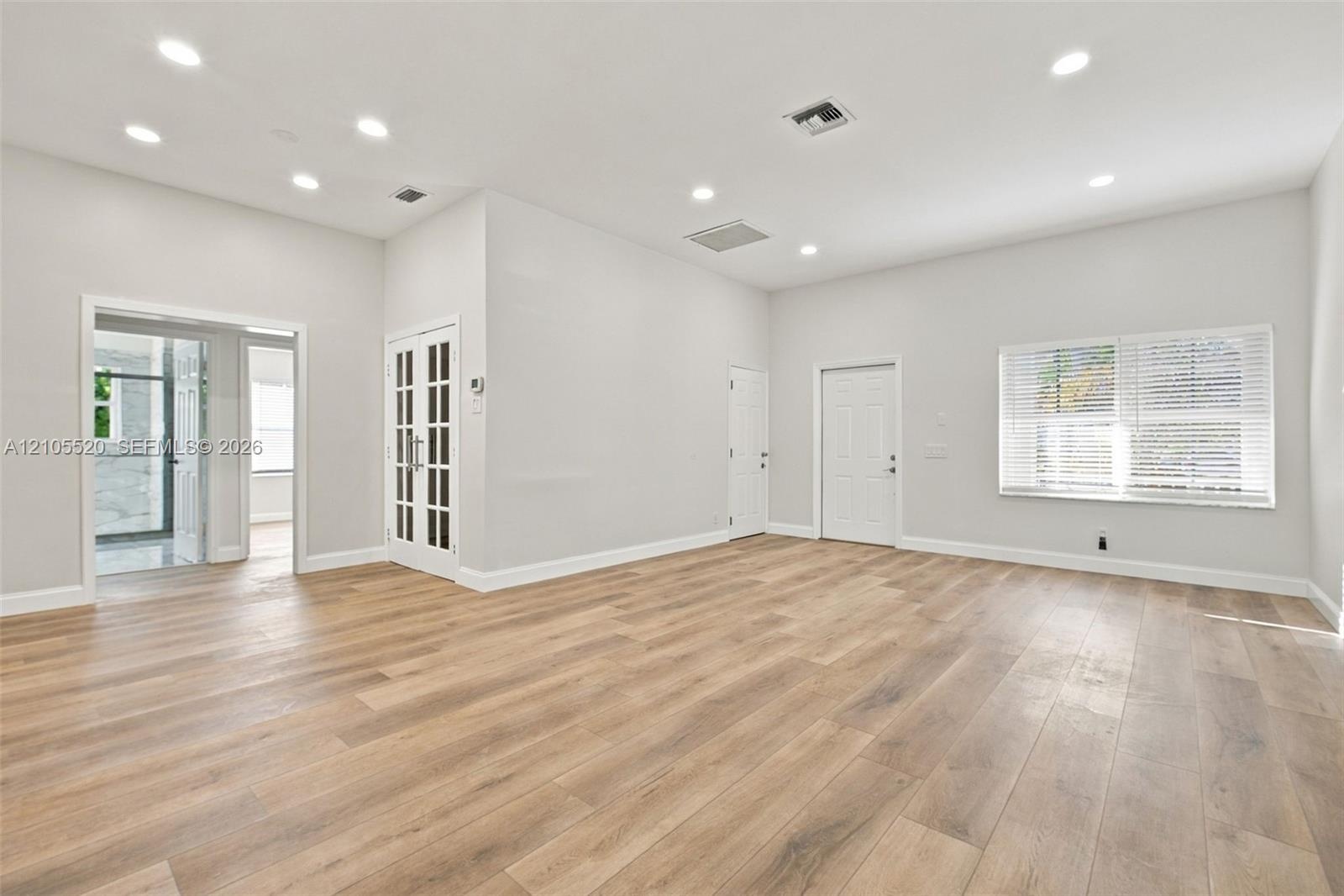 Open-concept room featuring wood-finish flooring, recessed lighting, white French doors with glass panels, and large windows with horizontal blinds