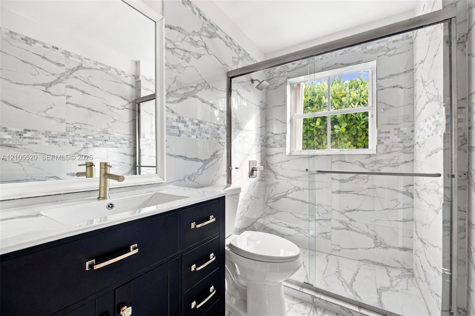 Dark navy vanity cabinet with metallic hardware, white countertop, large-format veined tile wall surfacing, glass-enclosed shower, and framed vanity m