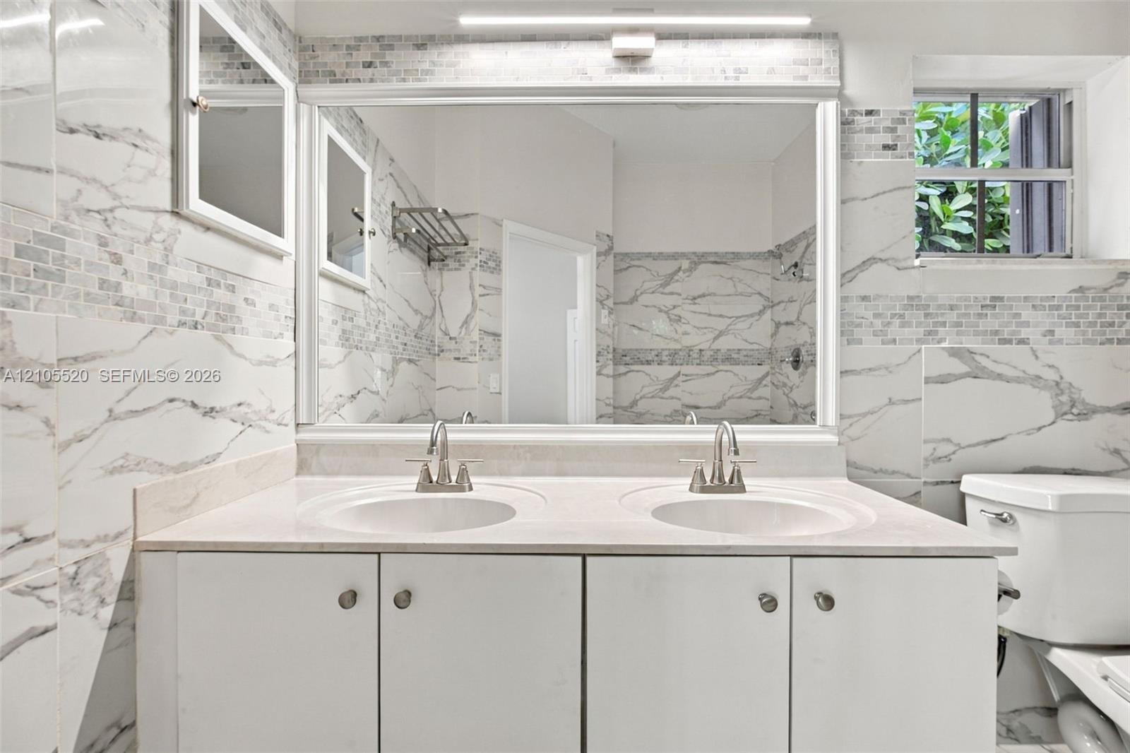 Double vanity featuring a stone countertop, dual undermount sinks, large rectangular mirror, and full-height stone-finish wall tile with accent inlay