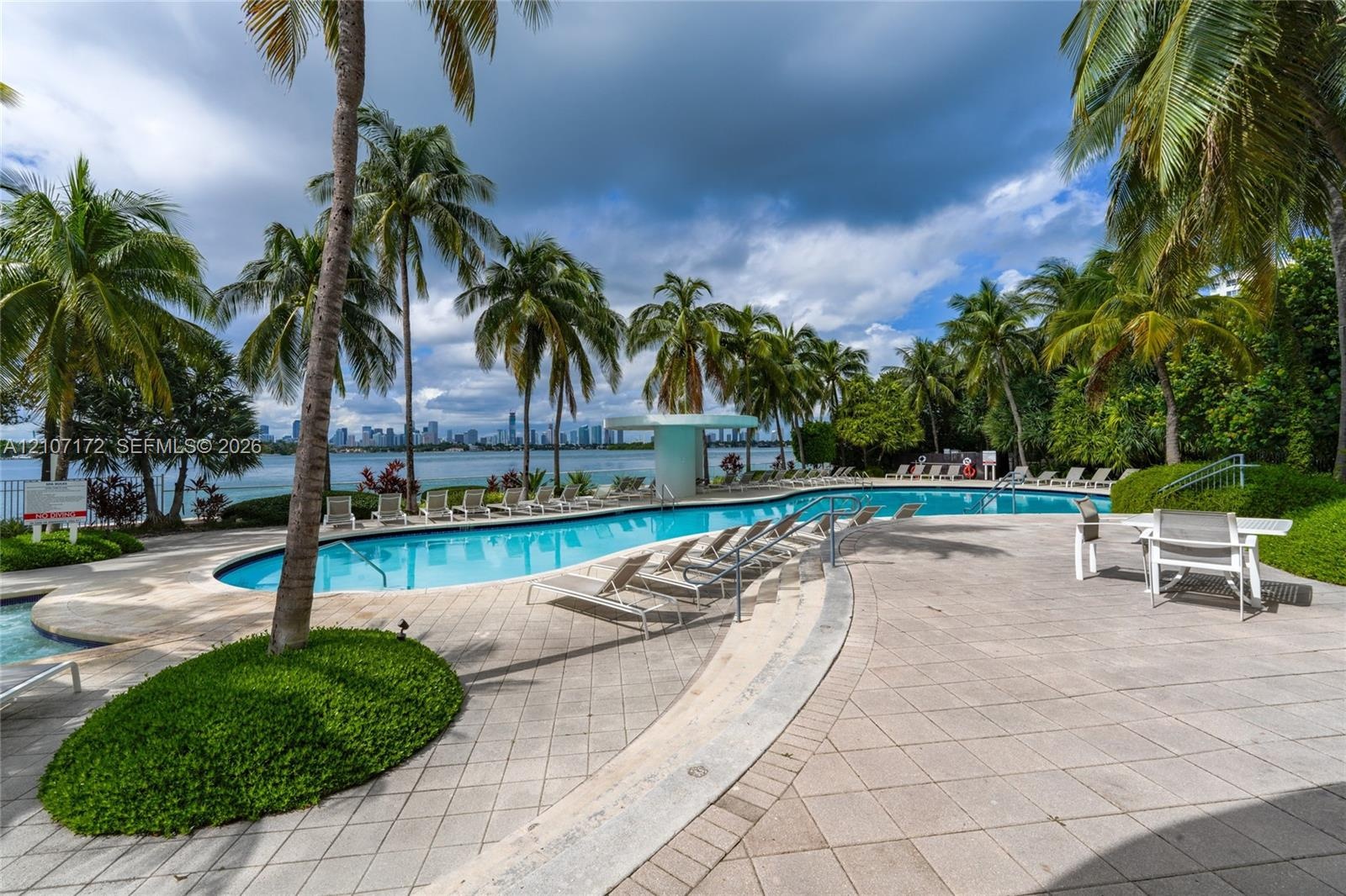 Outdoor patio featuring paved tile flooring, mature palm trees, string lighting, and an in-ground swimming pool overlooking the water