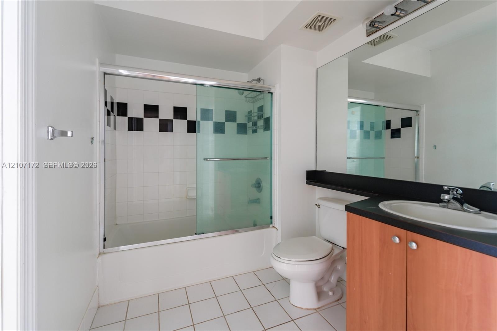 Bathroom featuring a vanity with wood-finish cabinetry, a black countertop with an integrated oval sink, a wall-to-wall mirror, and a shower-tub combi