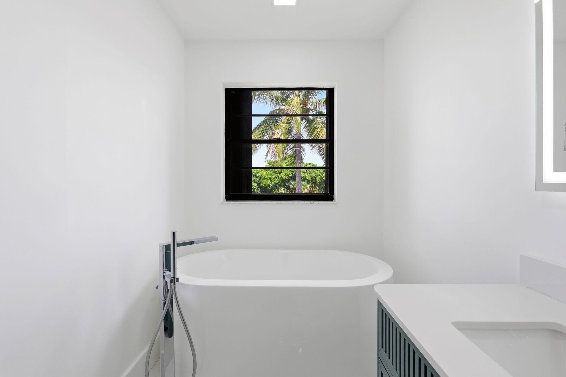 Freestanding soaking tub with floor-mounted chrome faucet, black-framed window, vanity with integrated sink, and illuminated mirror