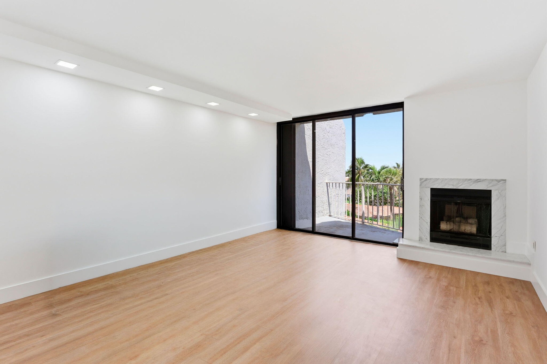 Spacious room with wood-finish flooring, a white marble fireplace, recessed ceiling lighting, and black framed glass sliding doors leading to a balcon