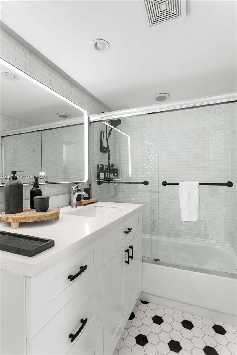 White shaker-style vanity with a solid surface countertop, undermount sink, integrated LED-backlit mirror, bathtub with glass sliding doors, and hexag