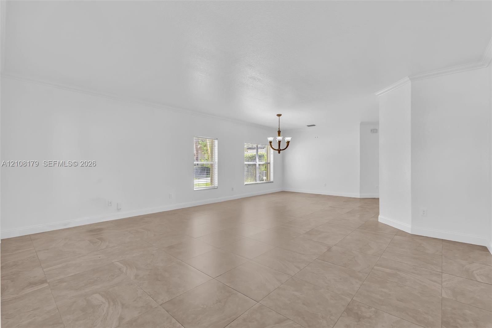 Open-concept interior featuring large-format tile flooring, recessed crown molding, dual windows, and a multi-arm chandelier