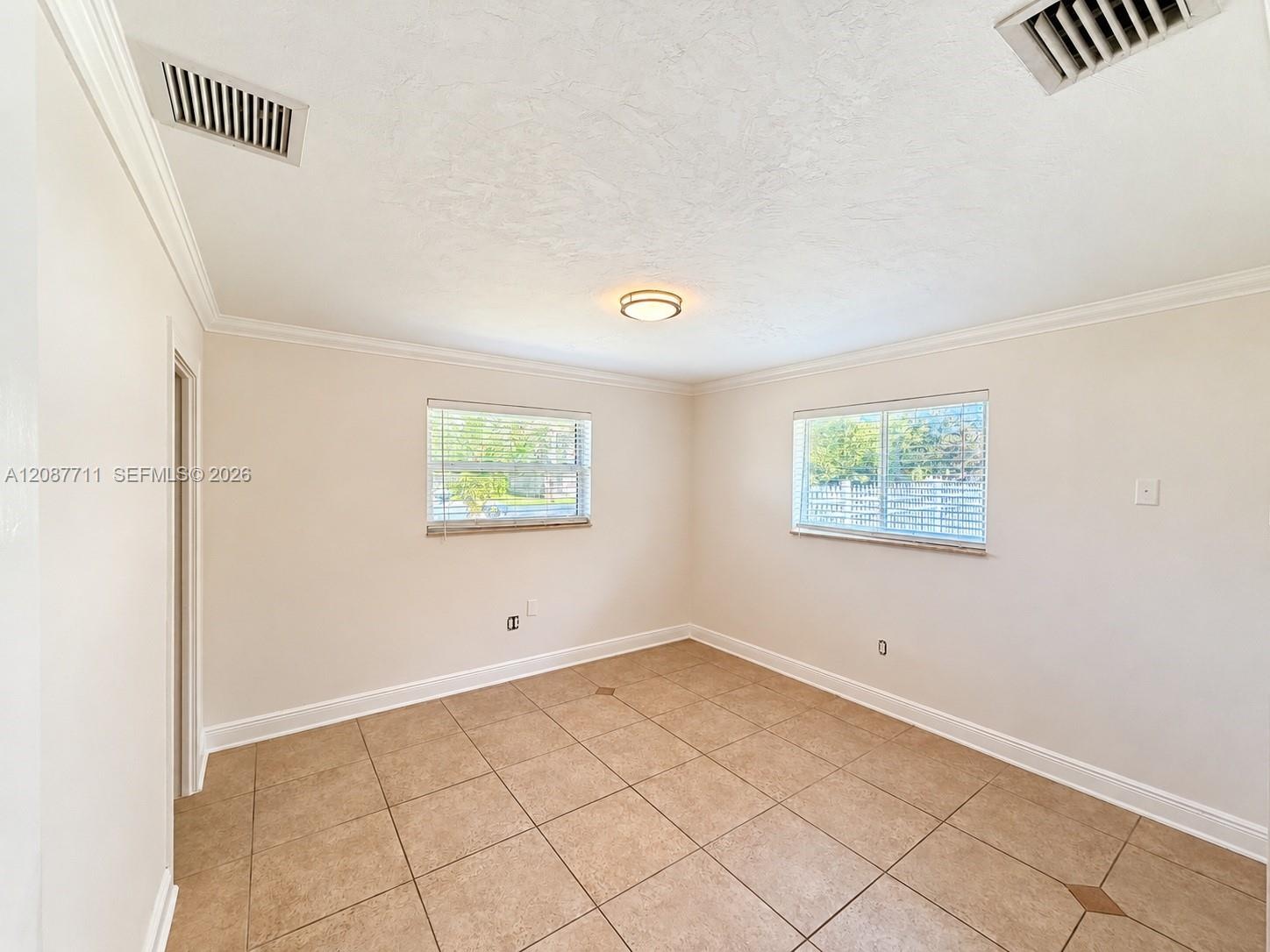 Neutral-toned interior featuring large-format square tile flooring, crown molding, dual windows with horizontal blinds, and a centered flush-mount cei