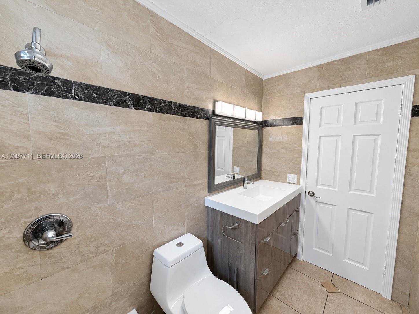 Full-wall tile surround with dark stone accent banding, wood-finish vanity with integrated sink, four-light vanity fixture, and six-panel interior doo