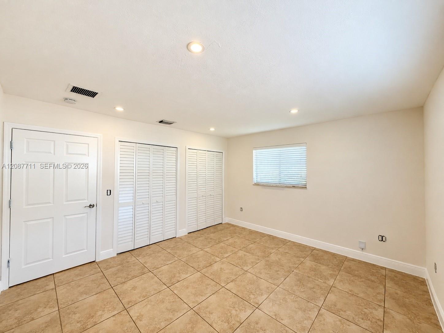 Room featuring neutral tile flooring, recessed ceiling lighting, two sets of white louvered-style closet doors, and a single window with integrated bl