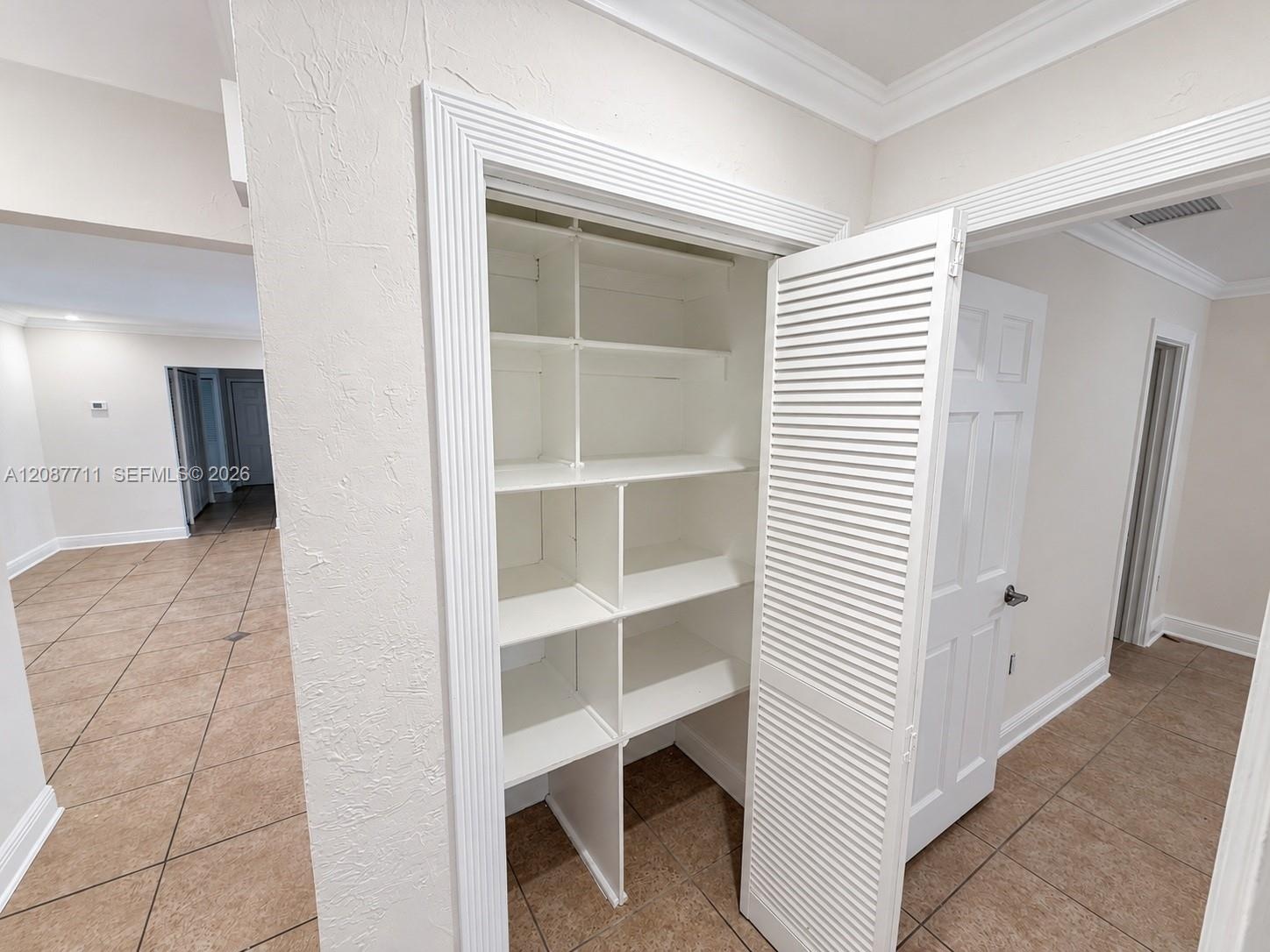 Built-in storage closet featuring custom shelving, louvered white door, decorative door casing, and tile flooring