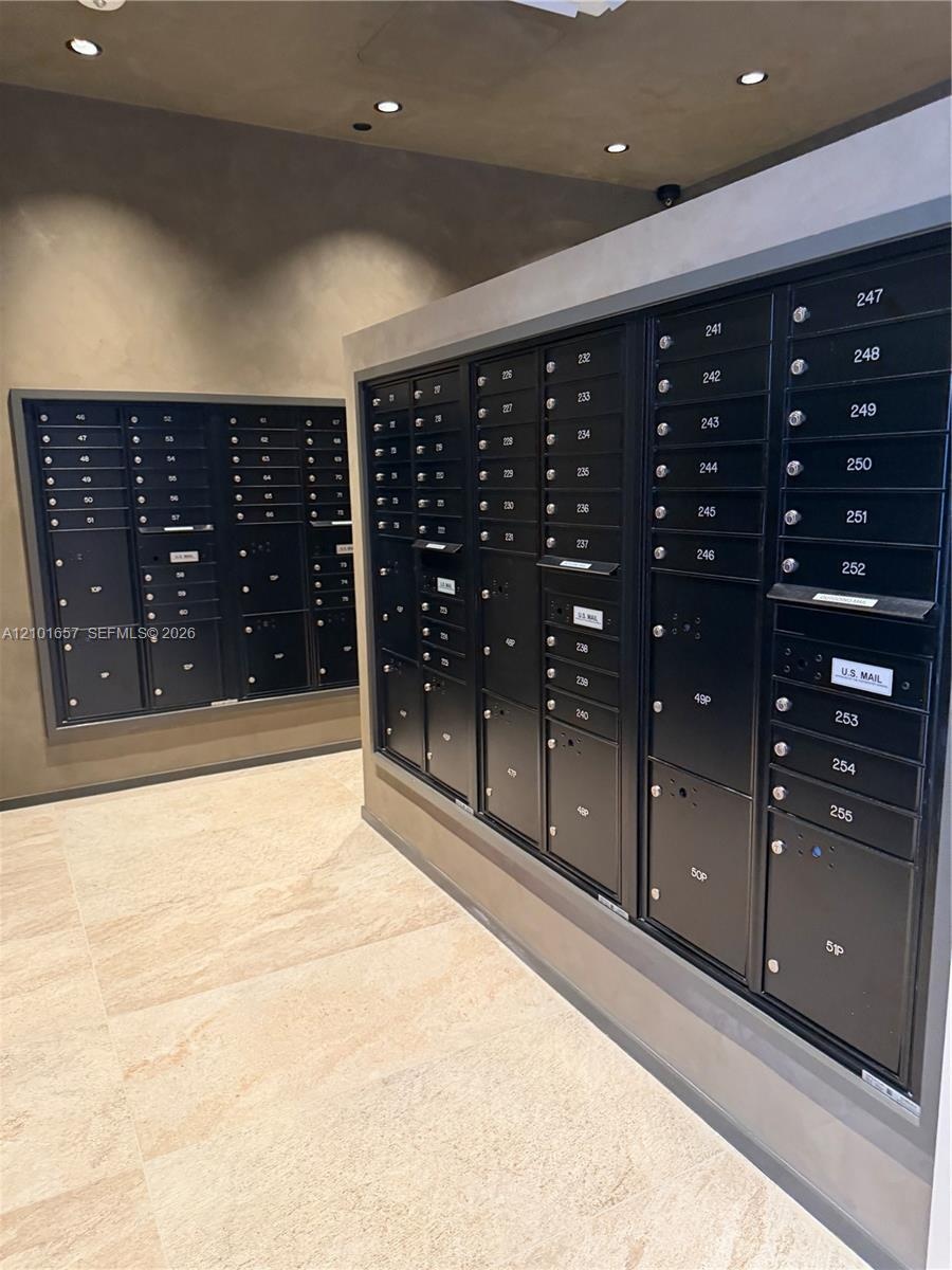 Integrated mail station featuring matte black secure postal lockers, neutral stone-finish tile flooring, recessed ceiling lighting, and smooth, monoch