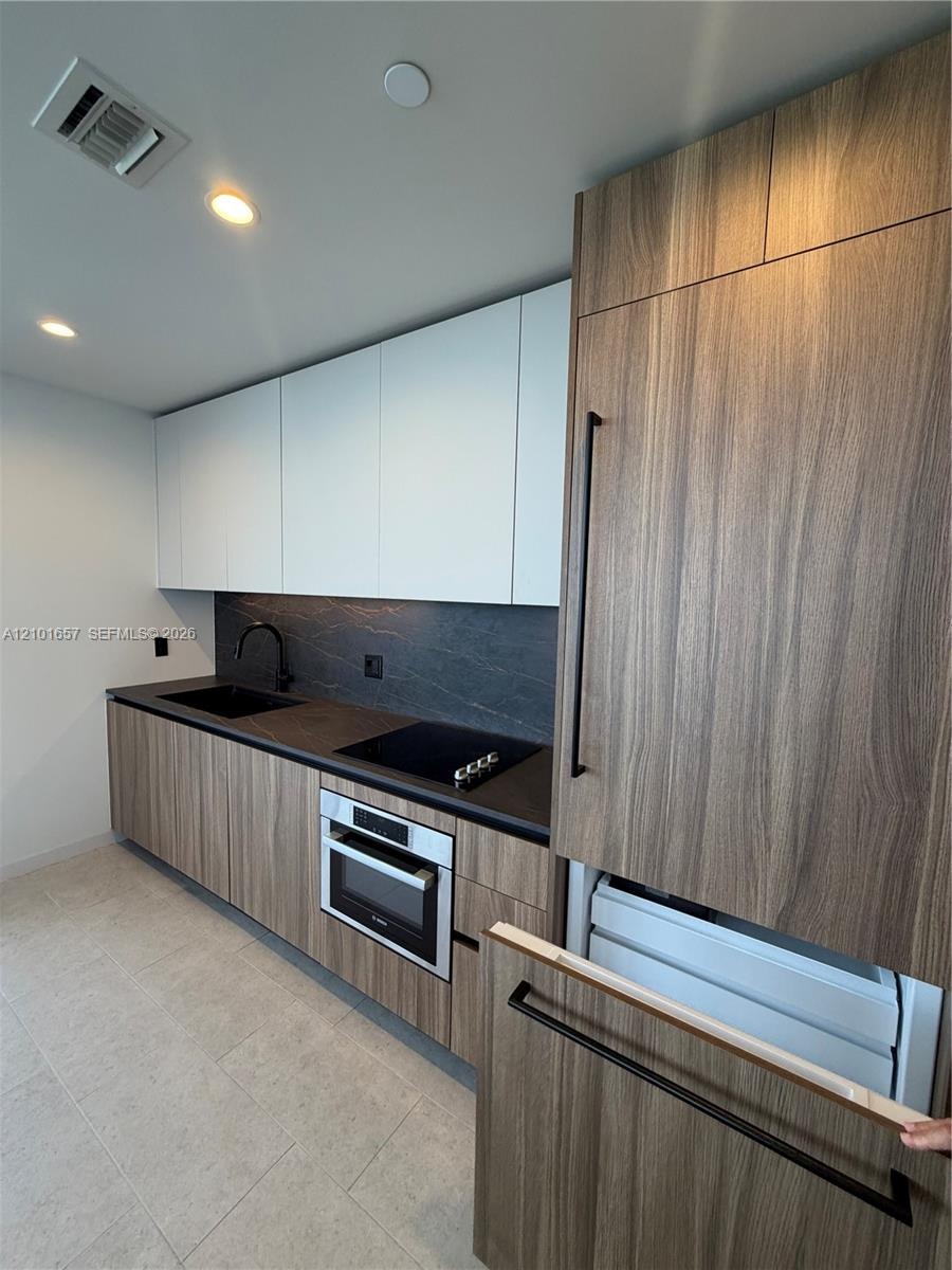 Kitchen featuring wood-finish cabinetry, matte black hardware, integrated appliance paneling, dark stone-finish countertops, and a stone-finish backsp