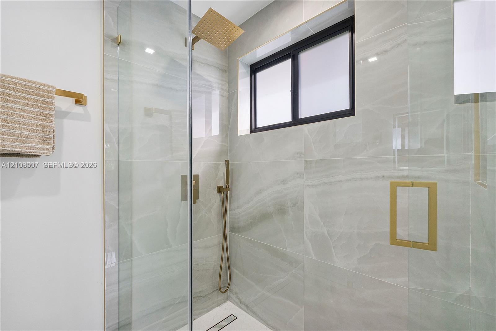 Walk-in shower featuring floor-to-ceiling stone-finish tile, glass enclosure, gold-tone hardware, rainfall showerhead, and a rectangular niche
