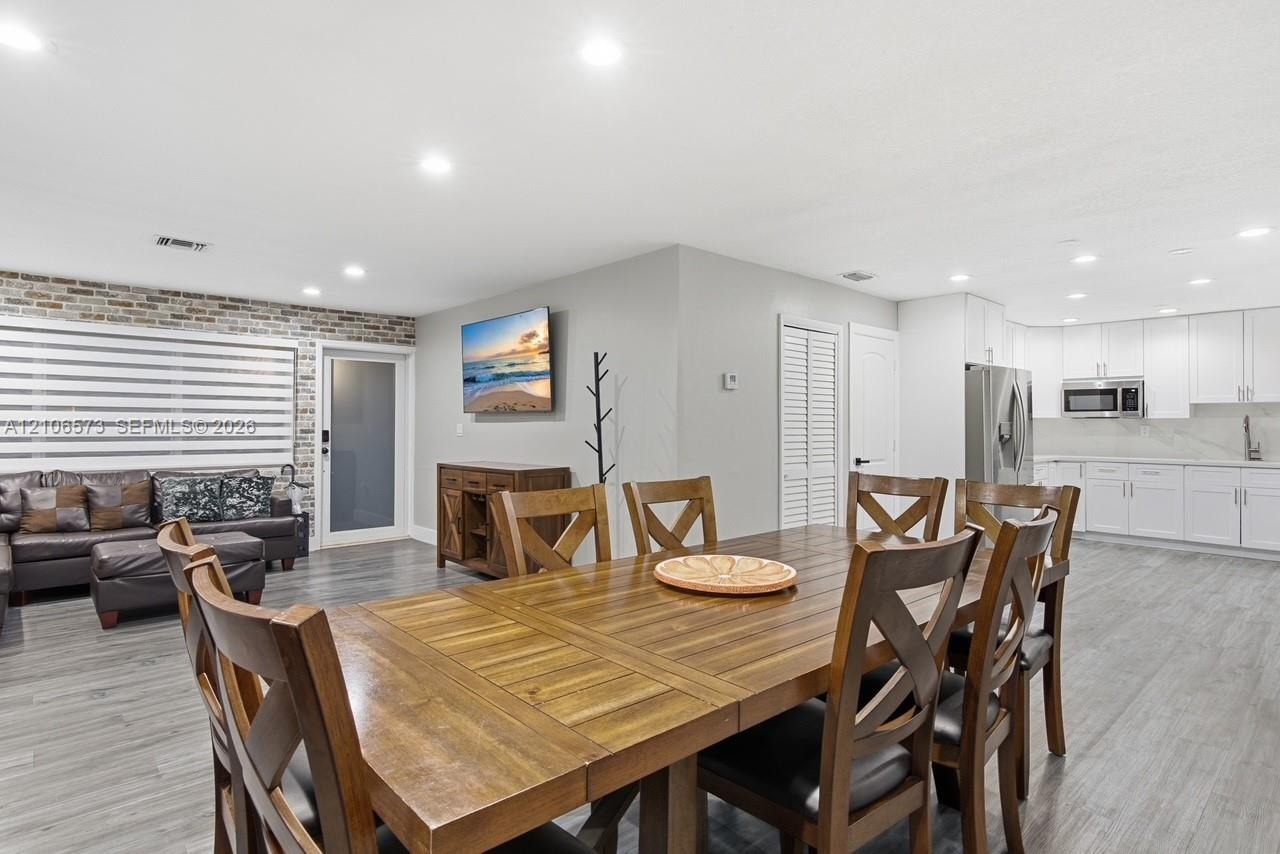 Open-concept living space featuring wood-finish flooring, recessed lighting, exposed brick accent wall, and a kitchen with white cabinetry and stainle