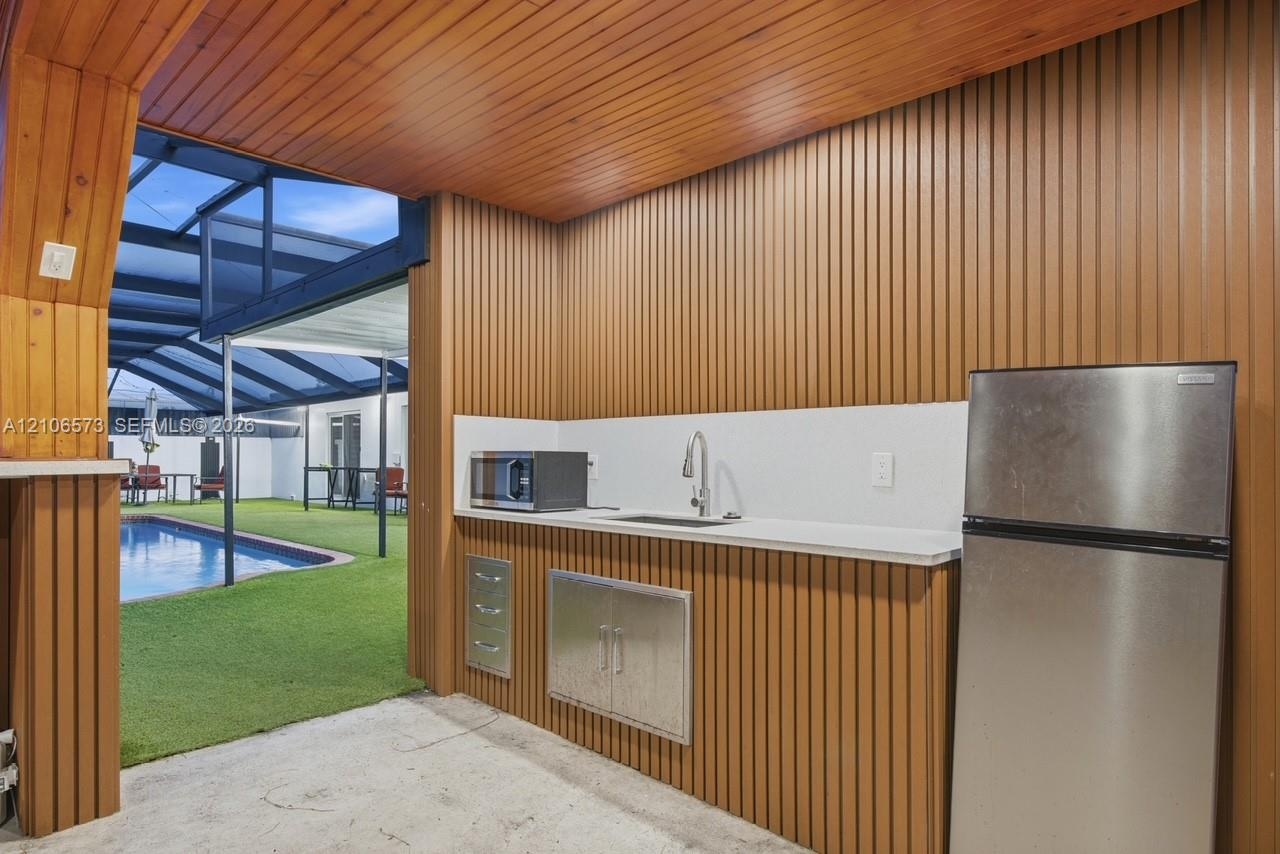 Covered outdoor kitchen featuring vertical wood-paneled walls and ceiling, stainless steel refrigerator, undermount sink with gooseneck faucet, and bu