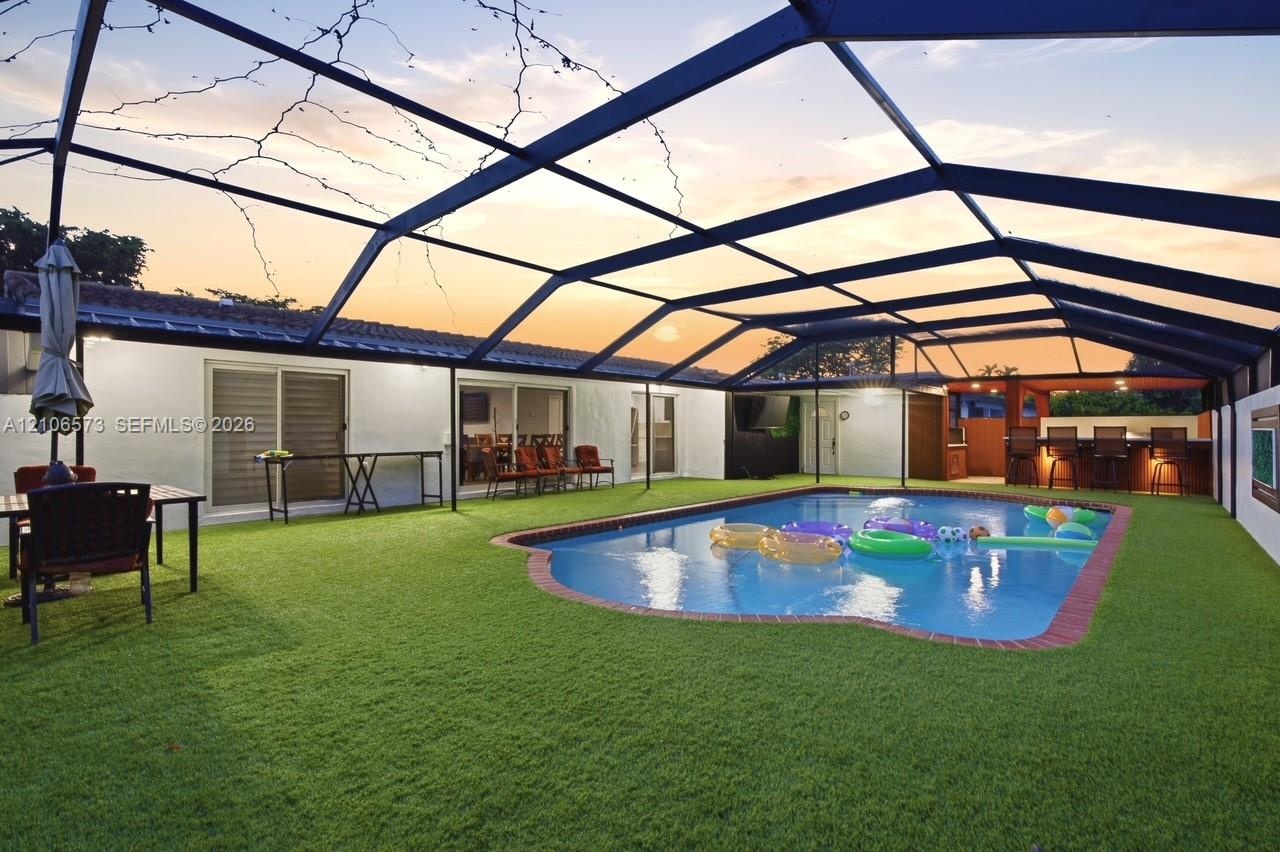 Screened-in outdoor living space featuring an in-ground swimming pool with brick coping, synthetic turf lawn, and a built-in outdoor bar