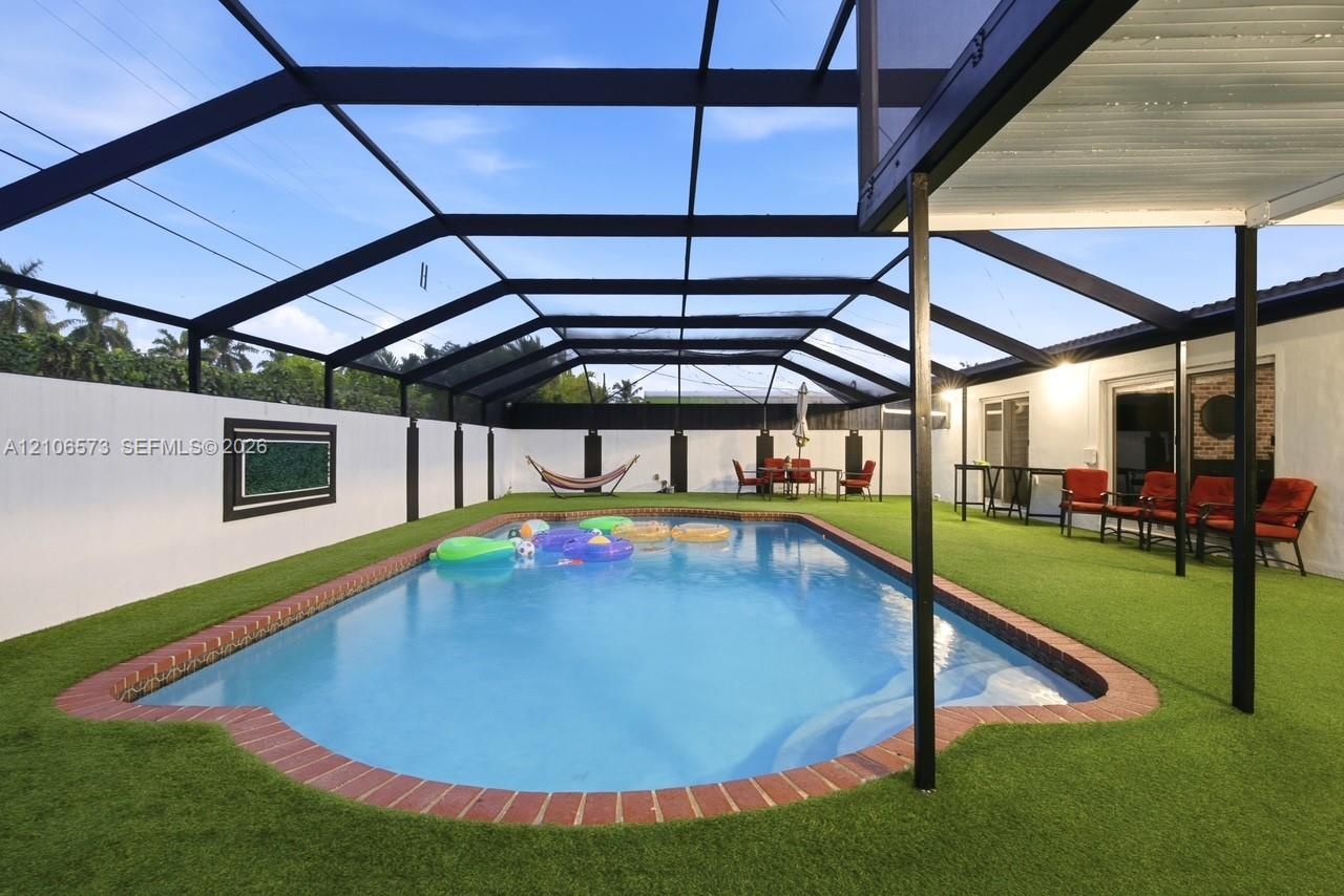 In-ground pool with brick coping, enclosed by a screened lanai structure, featuring artificial turf surfacing and a perimeter privacy fence