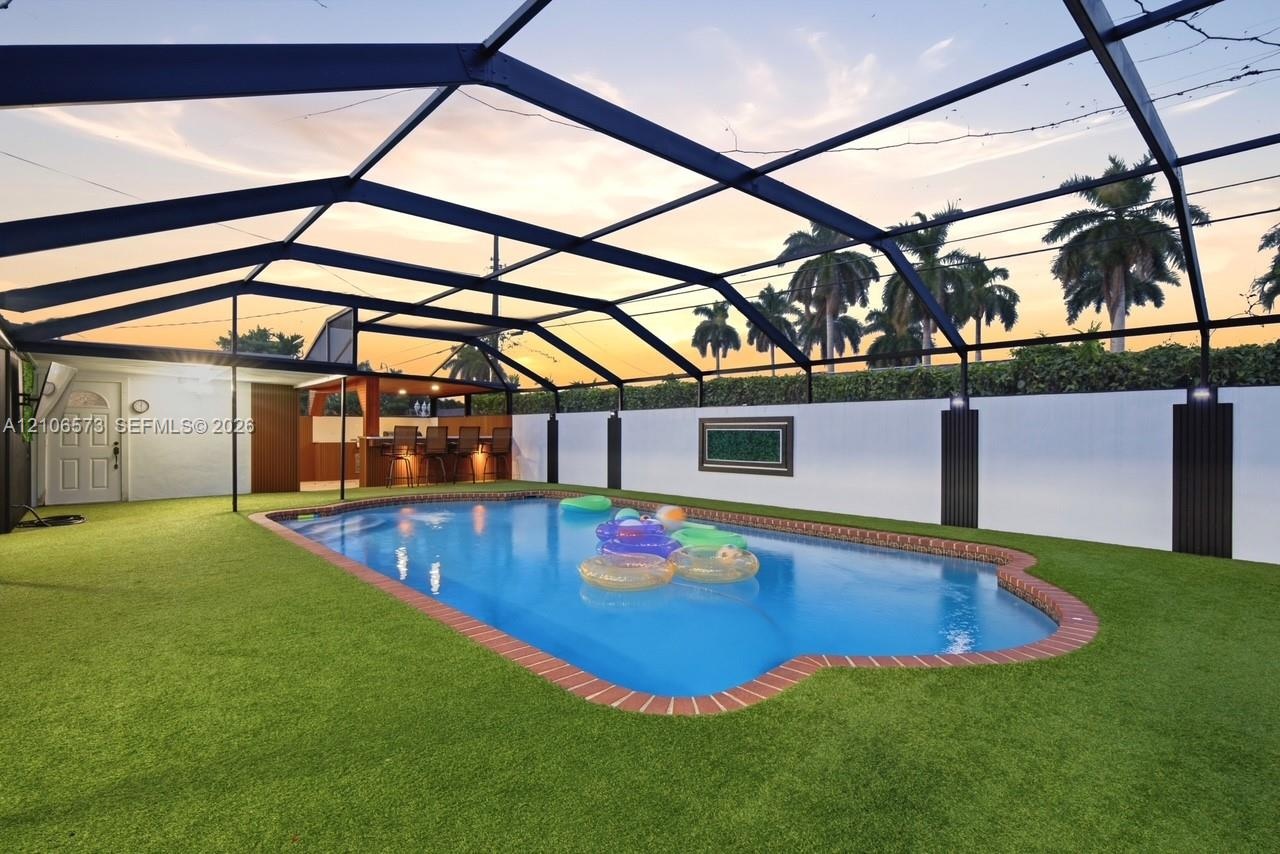 In-ground swimming pool with brick paver coping, fully enclosed by a screened lanai structure, featuring a built-in outdoor bar and synthetic turf sur