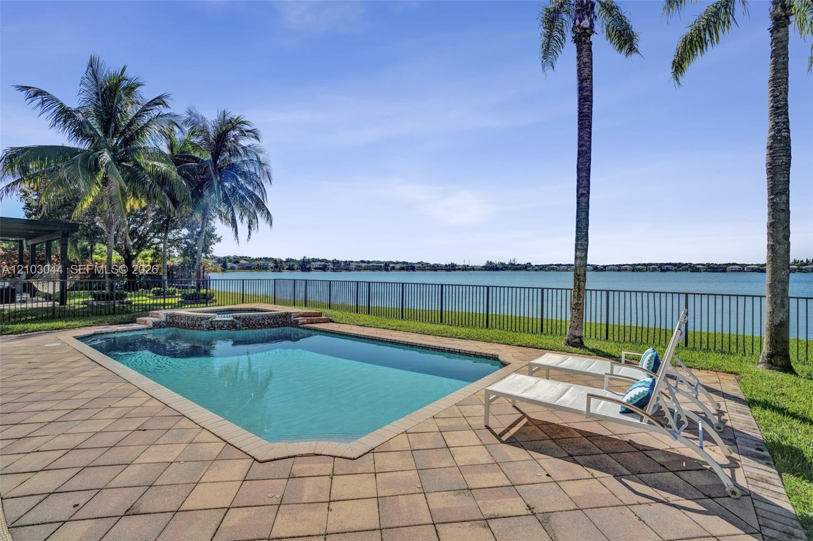 In-ground pool with stone-accented spillover spa, expansive paver patio, metal perimeter fencing, and mature palm trees overlooking the lake