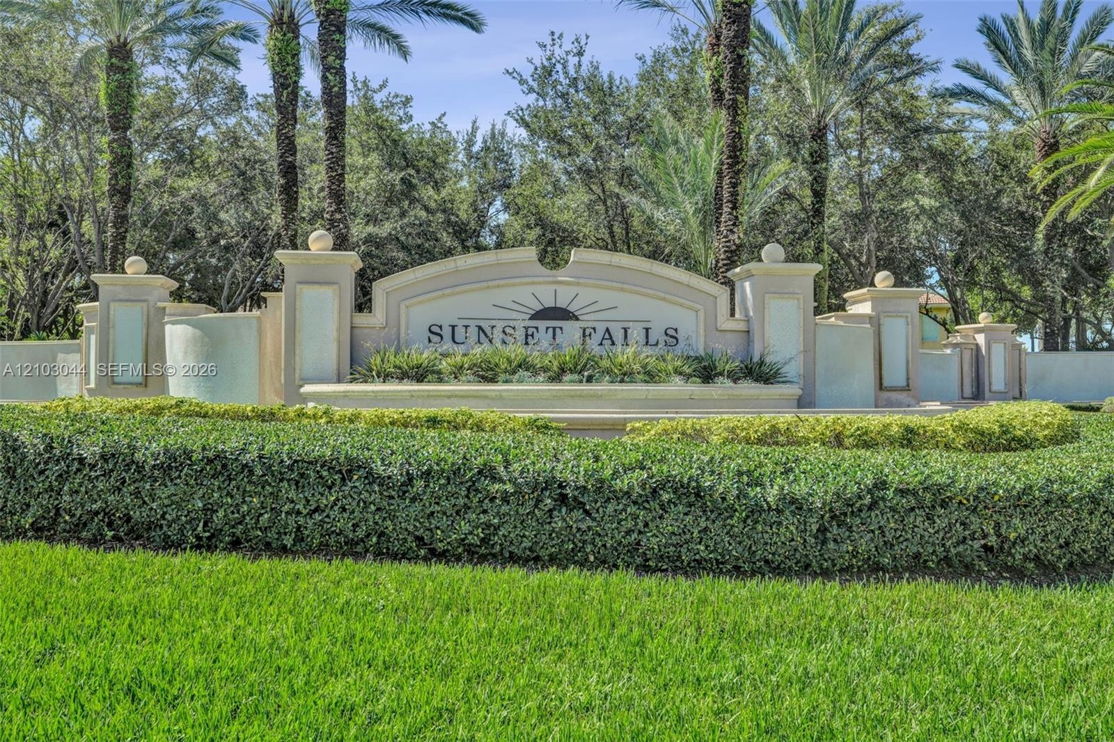 Stucco community signage featuring decorative masonry pillars, integrated lighting fixtures, tiered architectural detailing, and mature palm landscapi