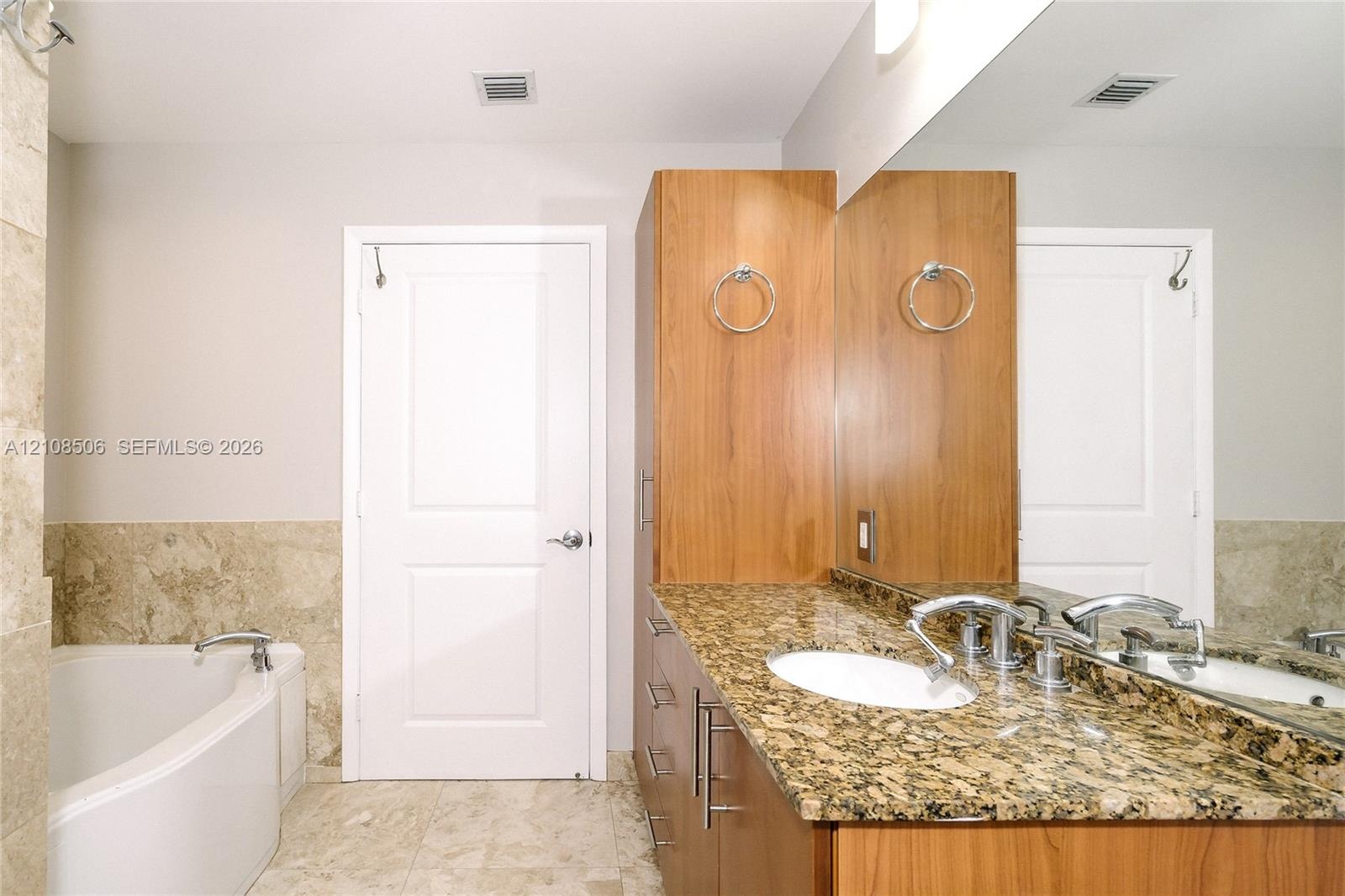 Bathroom featuring stone-surface vanity with dual integrated sinks, floor-to-ceiling wood cabinetry, large wall-mounted mirror, and a soaking tub with