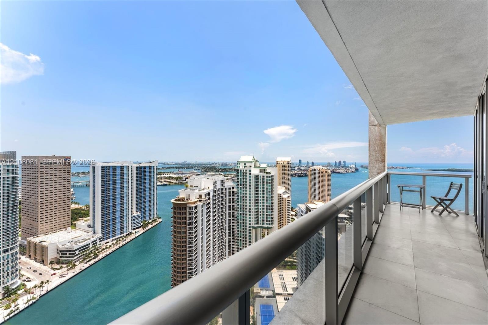 Covered balcony featuring large-format stone flooring and a glass-paneled railing overlooking the waterway and coastal skyline