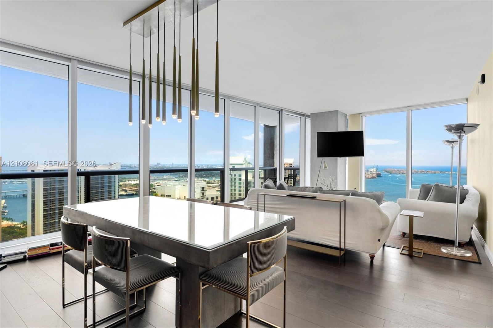 Floor-to-ceiling windows, wood-finish flooring, and a linear pendant light fixture featuring multiple metallic cylindrical elements