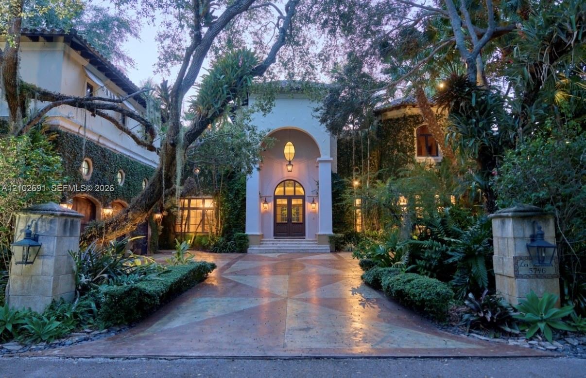 Stucco exterior residence featuring an arched portico entrance, geometric patterned driveway, stone entry pillars with mounted lanterns, and exterior 