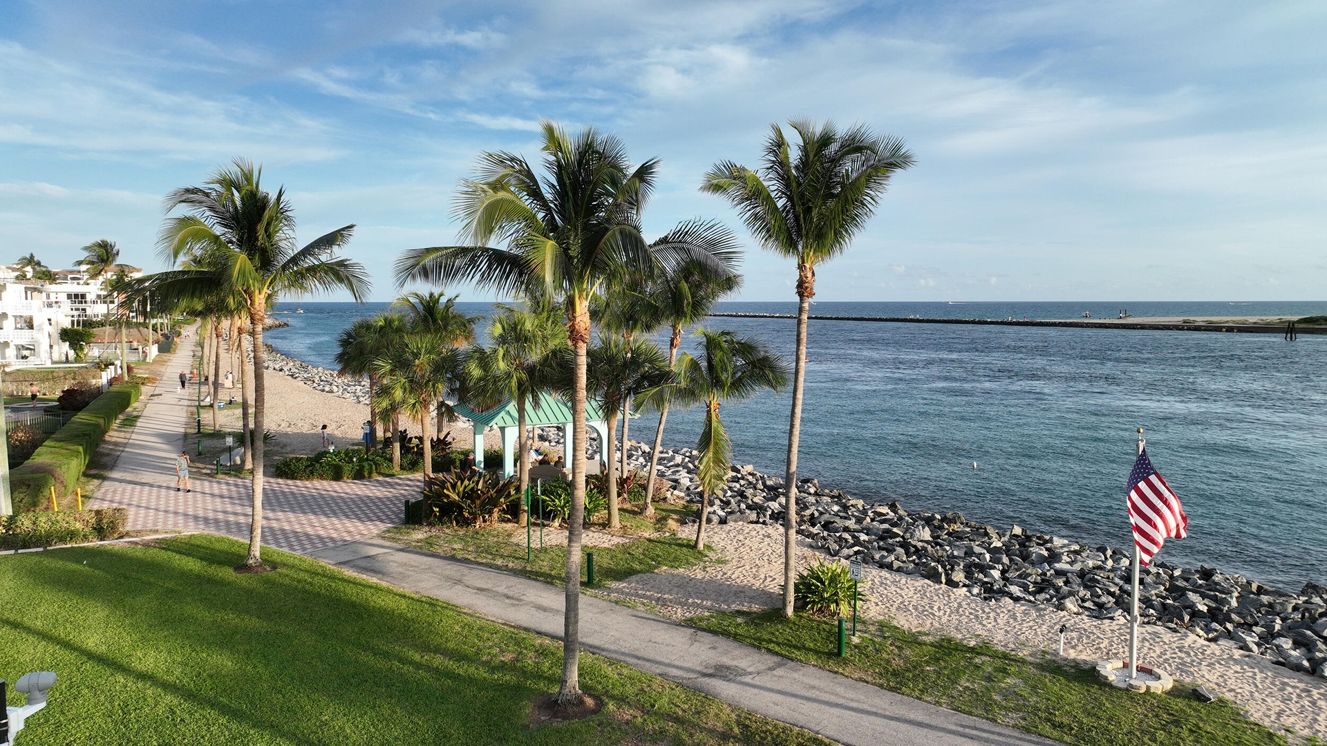 Waterfront property featuring a paved promenade, mature palm trees, expansive lawn, and a rock-lined shoreline