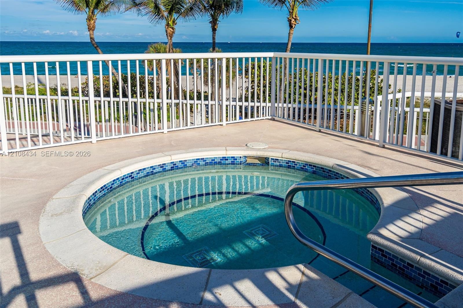 Circular jetted spa with blue tile surround, concrete decking, metal safety railing, and ocean backdrop