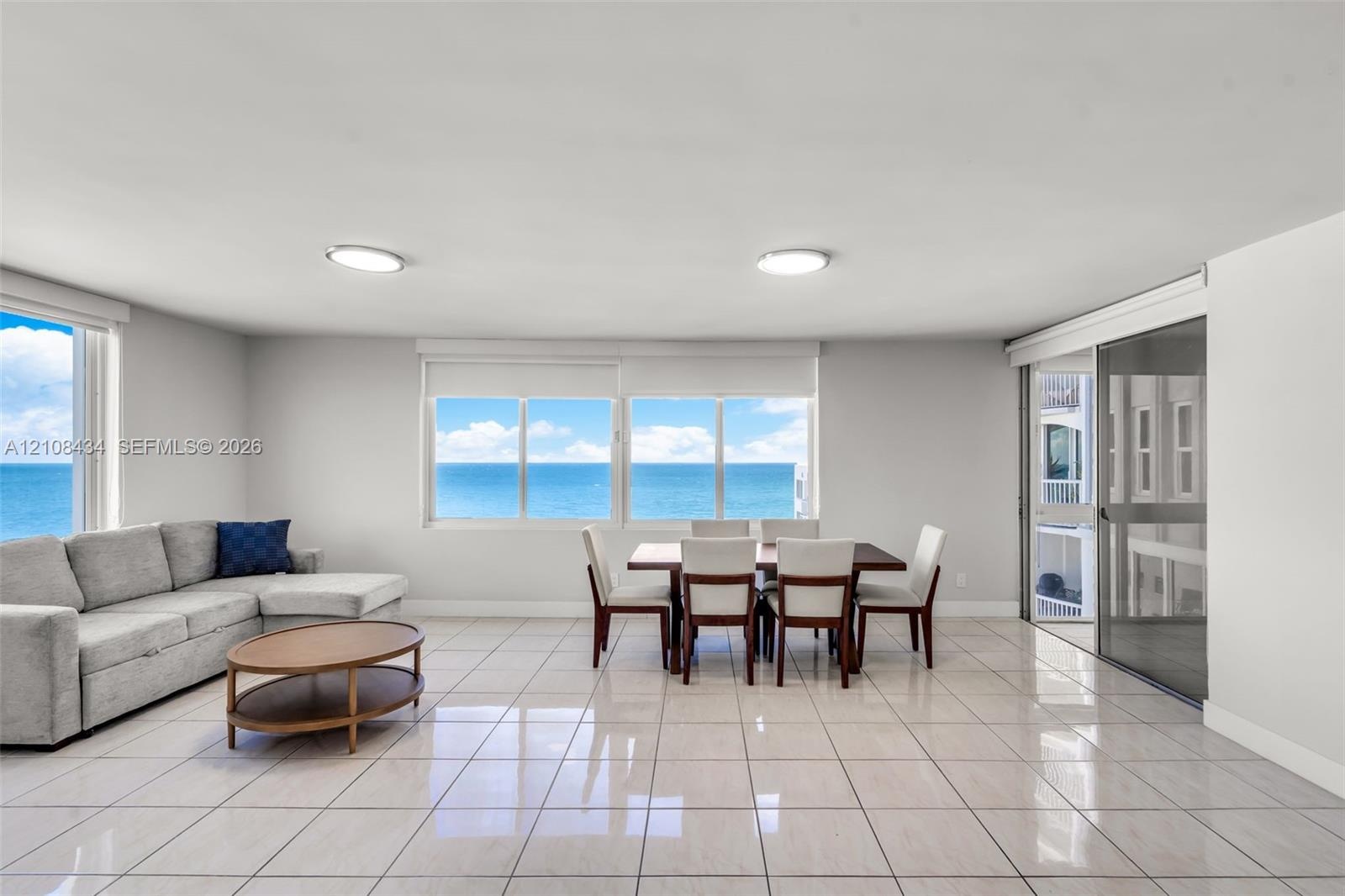 Open-concept room featuring light-toned tile flooring, large multi-pane windows, and a sliding glass door providing access to a balcony with an ocean 