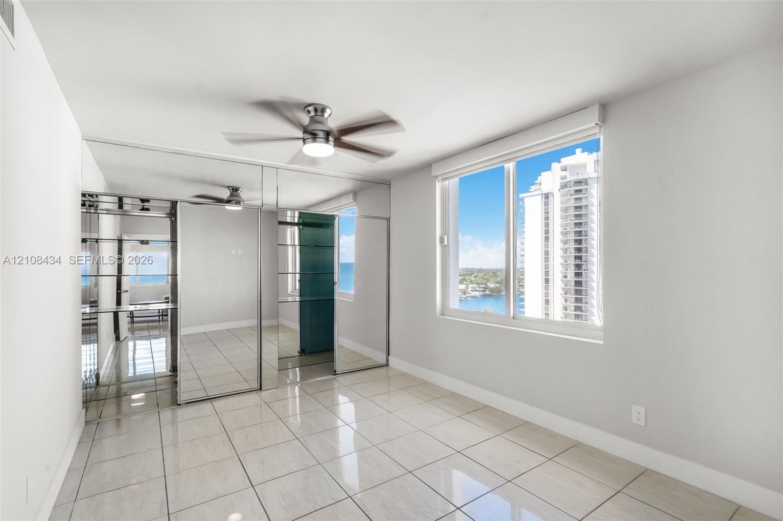 Large format tile flooring, floor-to-ceiling mirrored closet doors, ceiling fan with integrated lighting, and expansive window with water and skyline 