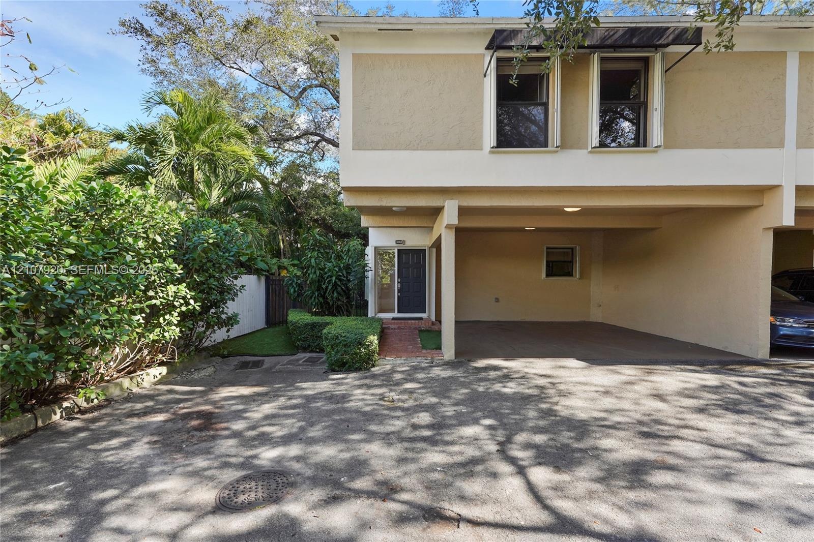Two-story exterior featuring a covered carport, brick paver entryway, dark-frame windows with metal awnings, and landscaped surroundings