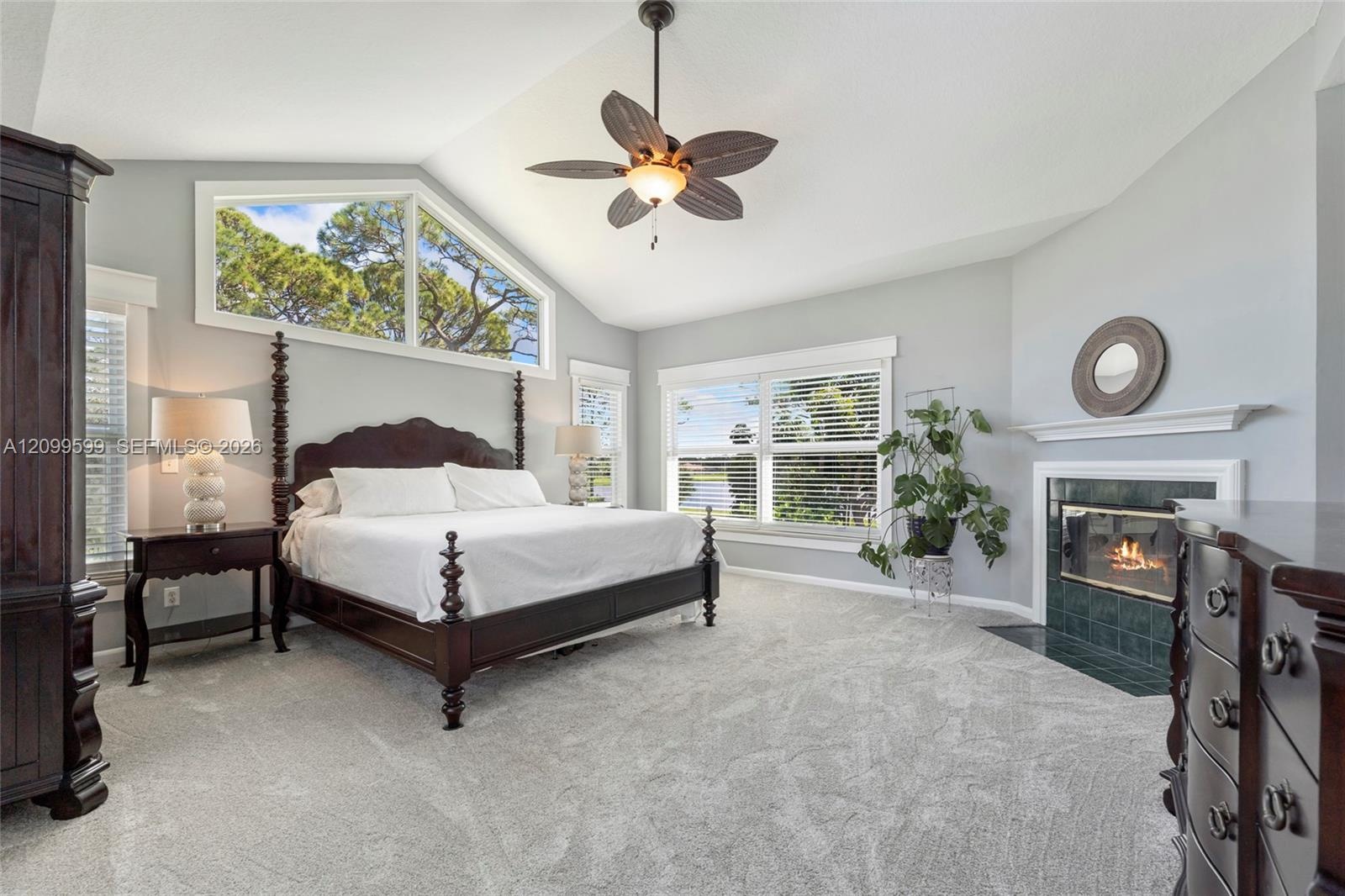Vaulted ceiling with a central multi-blade ceiling fan, expansive trapezoidal window, fireplace with a tiled hearth and mantel, and wall-to-wall carpe