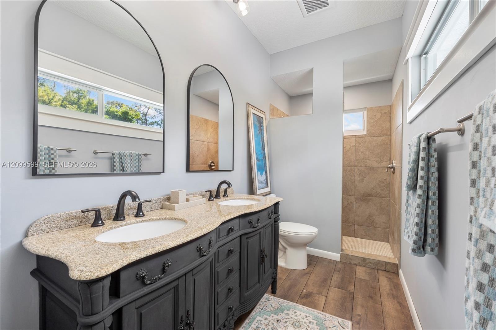 Dual-sink vanity with stone countertop and dark cabinetry, arch-top mirrors, wood-finish flooring, and a tiled shower enclosure